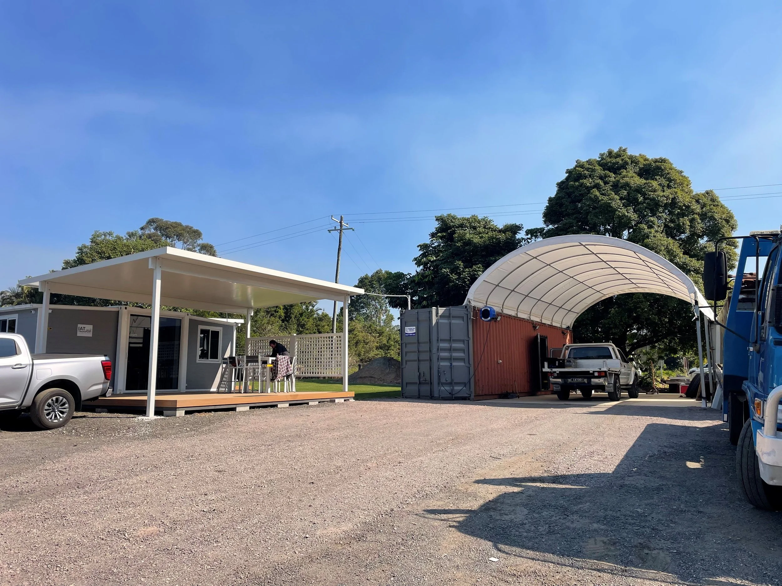 A construction site with a modular building, a covered storage area, and a white pickup truck parked on gravel under a clear blue sky. Trees and utility poles are visible in the background.