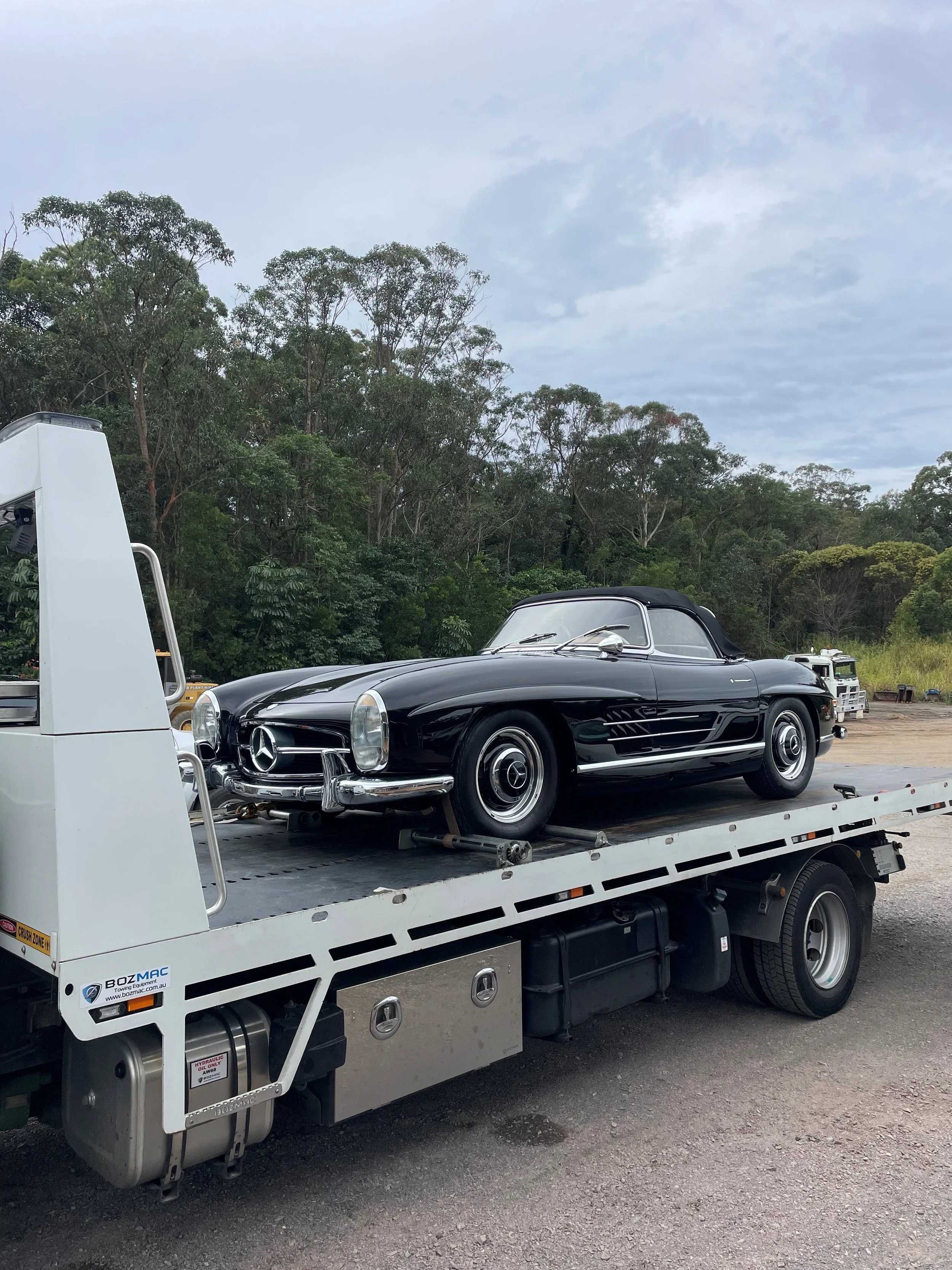 A vintage black Mercedes-Benz convertible on a flatbed tow truck in an outdoor area with trees in the background.