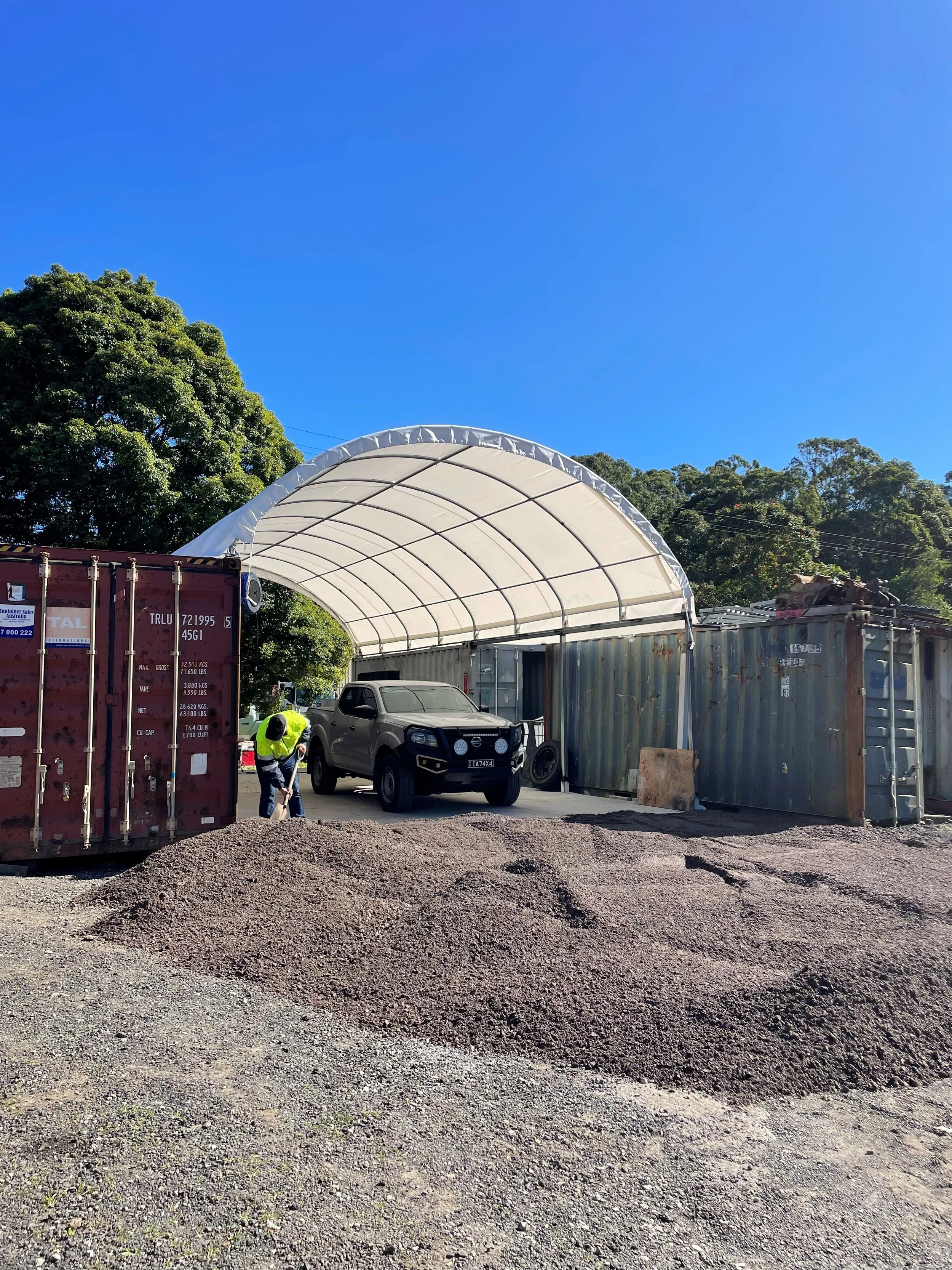 A construction site with a gravel ground, a black pickup truck parked under a white curved tent, a worker in a yellow safety vest, and several storage containers, with trees and a clear blue sky in the background.