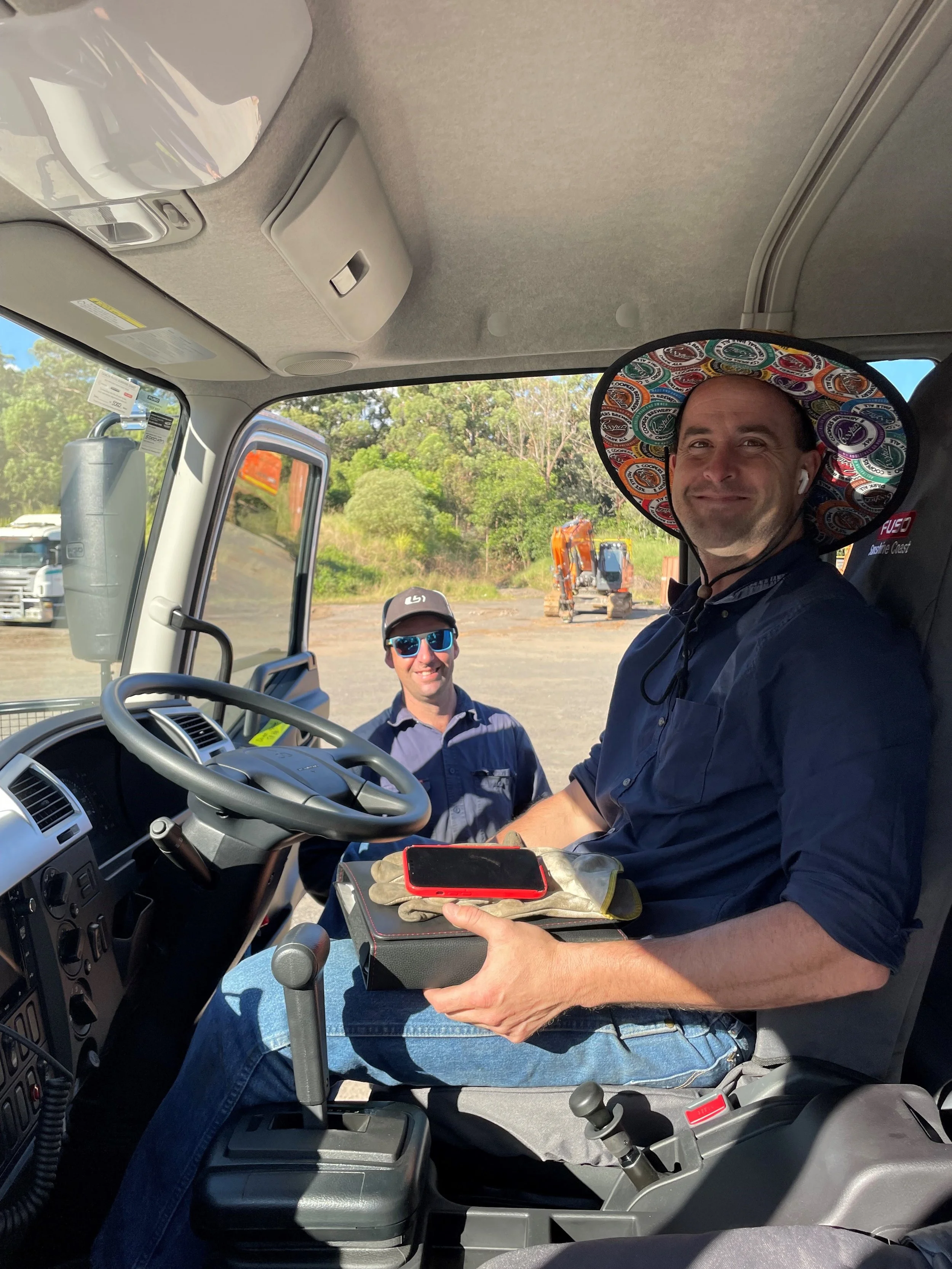 Two men in work uniforms, one sitting in the driver's seat of a truck wearing a colorful wide-brimmed hat, and another standing outside, both smiling and talking. The man in the truck is holding a glove, a red smartphone, and a box. Construction equipment and trees are visible outside.