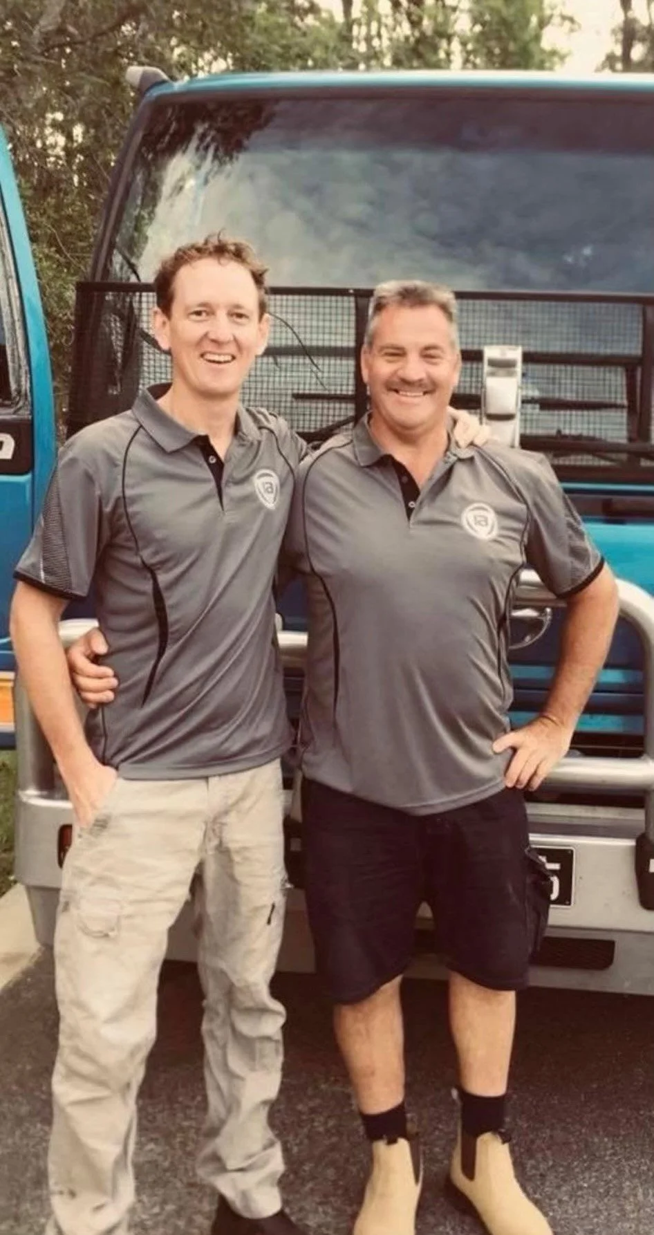 Two men wearing matching gray polo shirts with a logo, standing side by side in front of a truck. They are smiling and have their arms around each other.