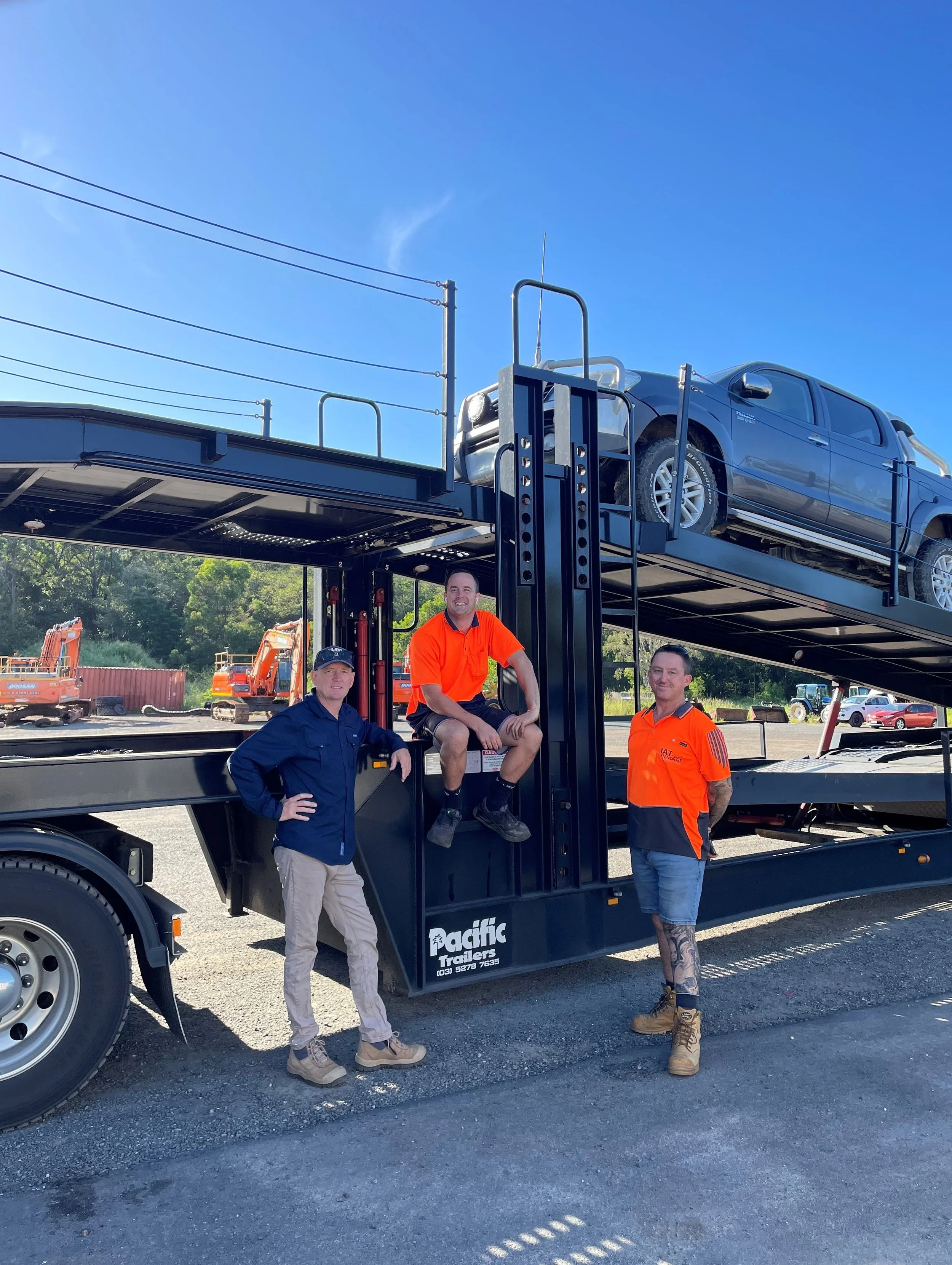 Three men standing and sitting in front of a black car carrier truck with a loaded car on top. The background shows construction equipment and trees under a clear blue sky.