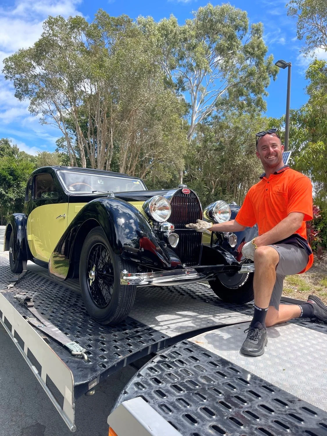 A man kneeling next to a vintage black and yellow car on a flatbed truck, smiling outdoors with trees and blue sky in the background.
