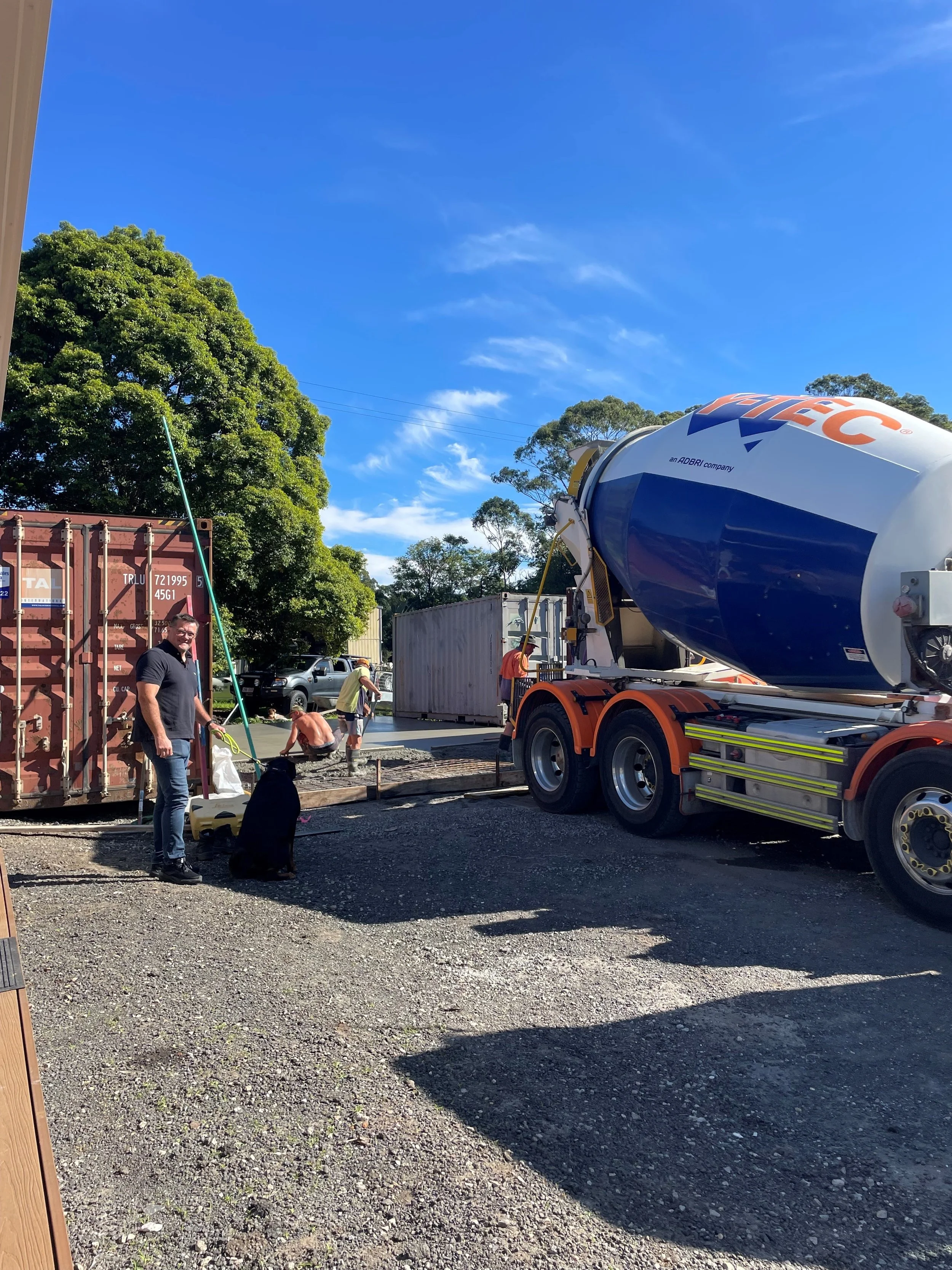 Construction site with workers pouring concrete, a cement mixer truck, and a man with a black dog, set under a clear blue sky with trees in the background.
