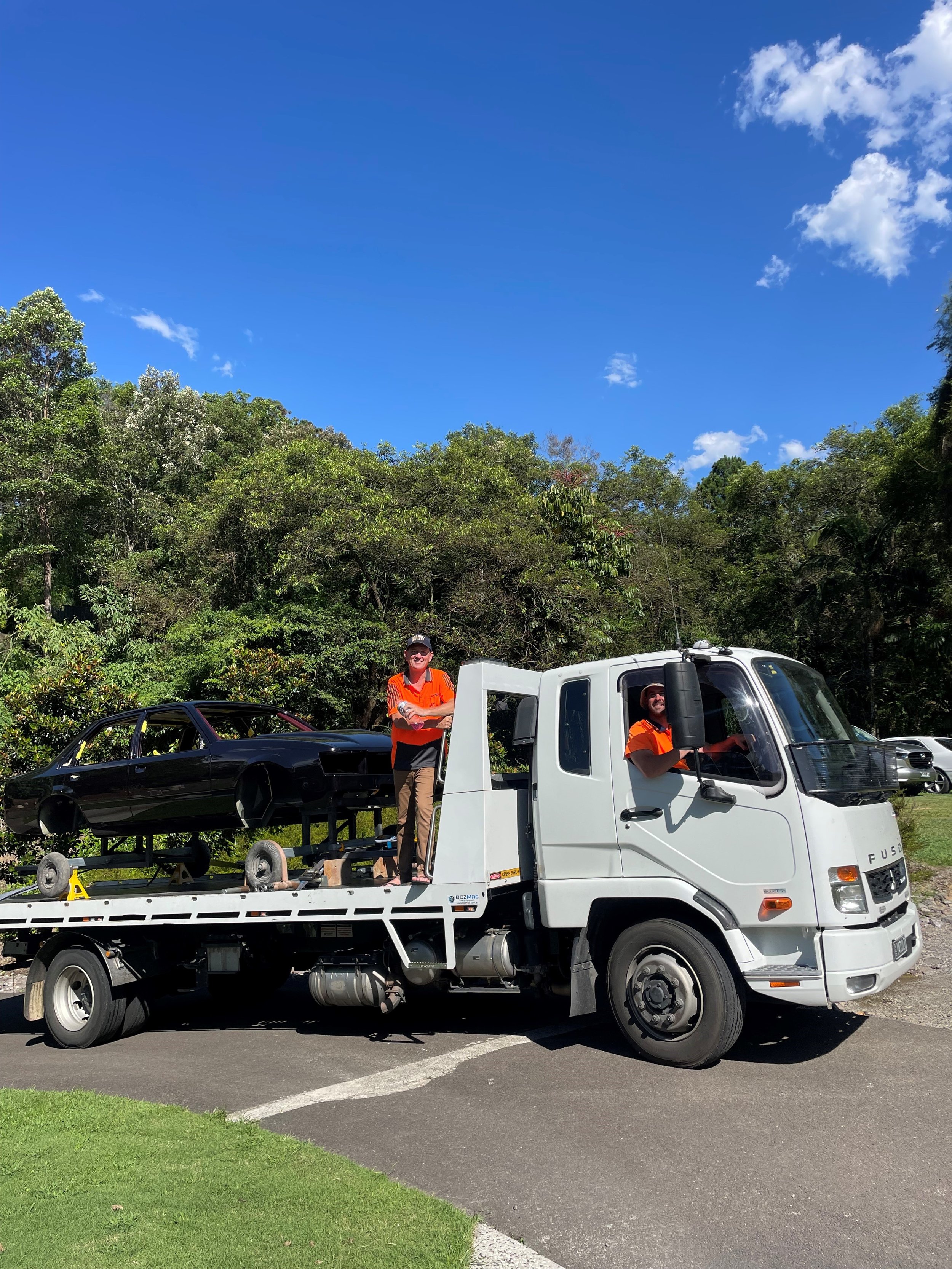A tow truck carrying a black car with no front end, parked on the side of a road. Two people in orange shirts are standing and sitting on the truck, smiling. The background features green trees and a bright blue sky with some clouds.