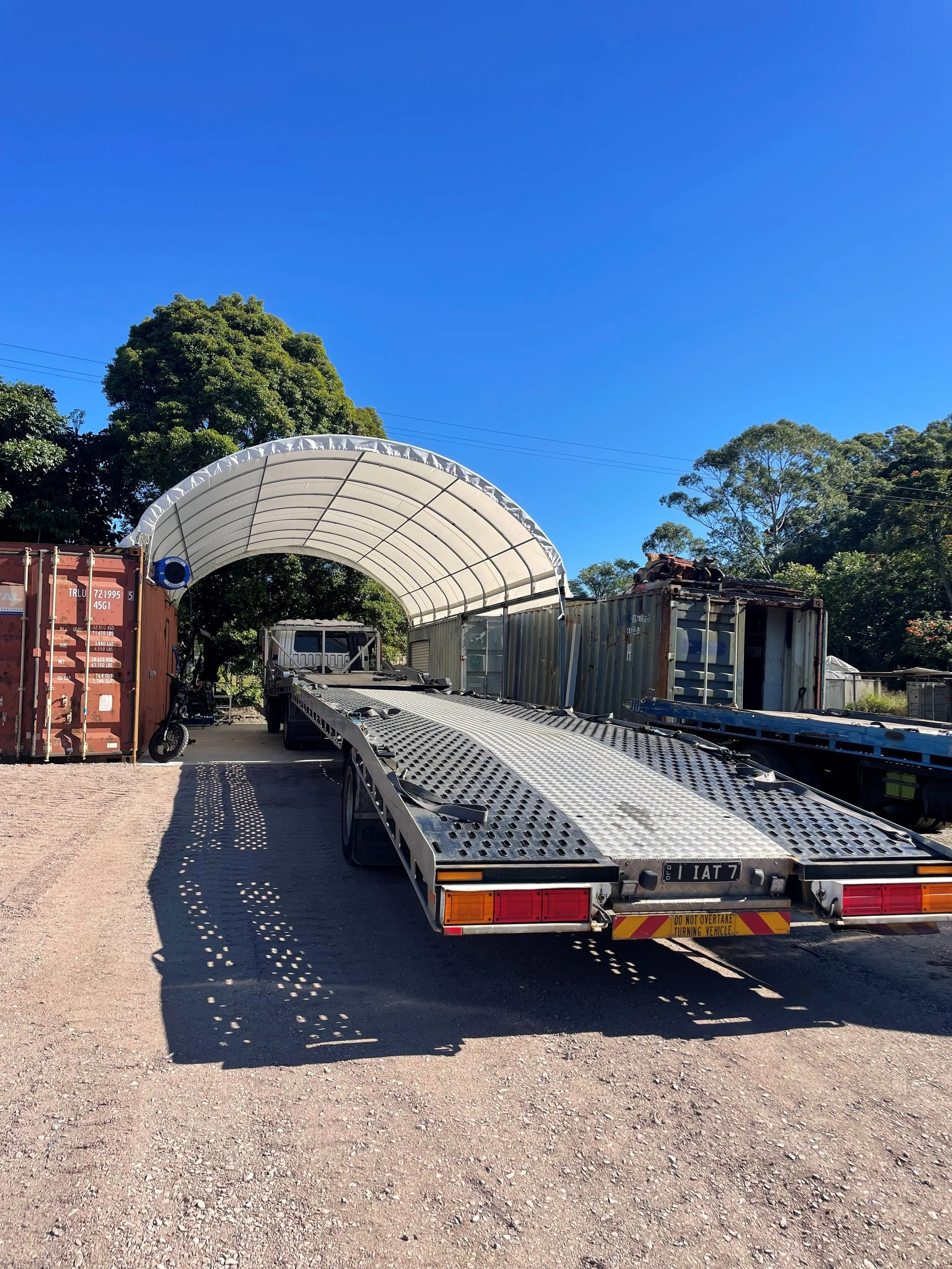 Flatbed trailer parked under a fabric canopy with shipping containers and trees in the background.