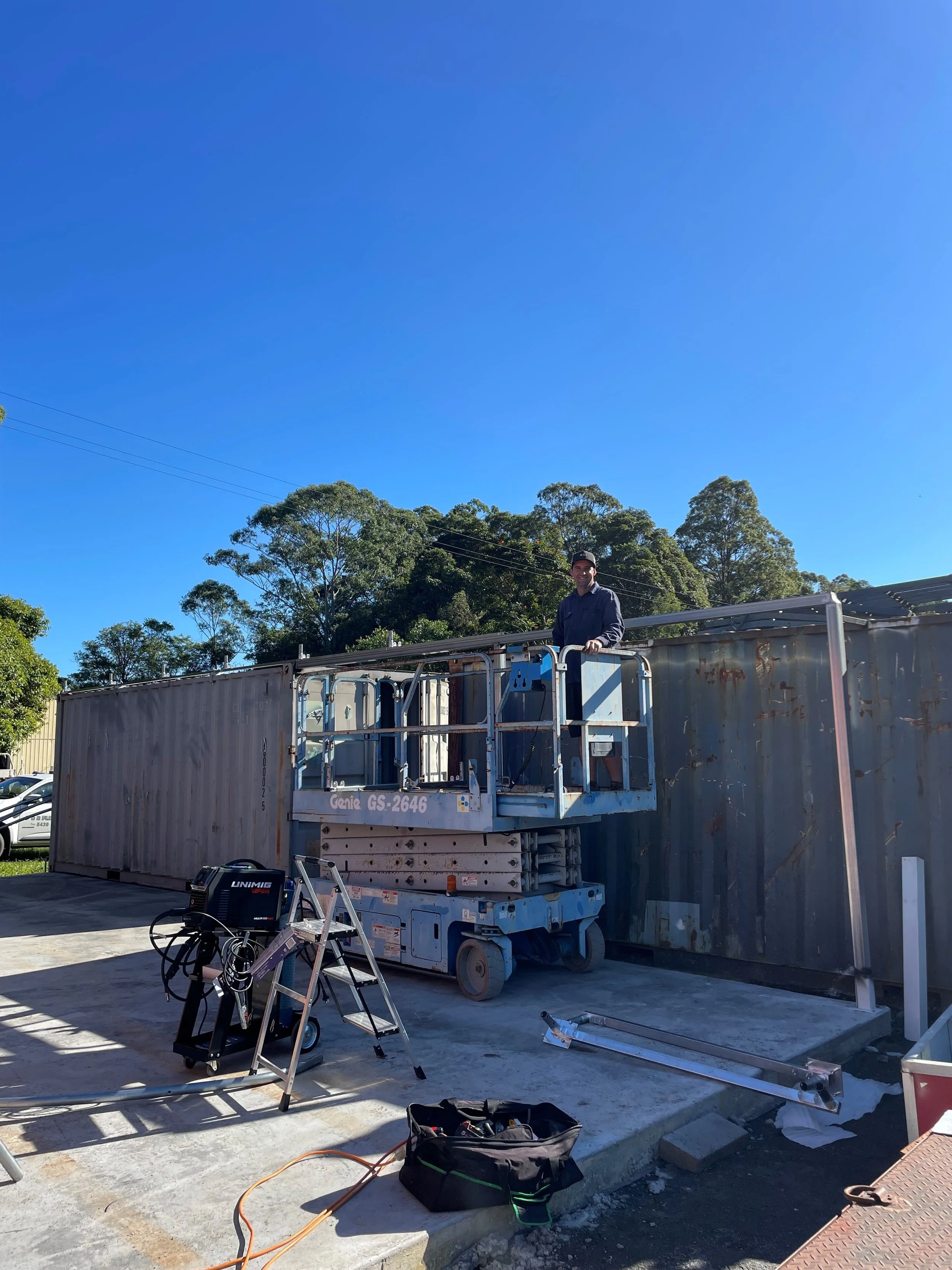 A man standing in a lift basket on a concrete surface next to a large rusty shipping container, with electrical equipment and tools nearby, under a clear blue sky and trees in the background.