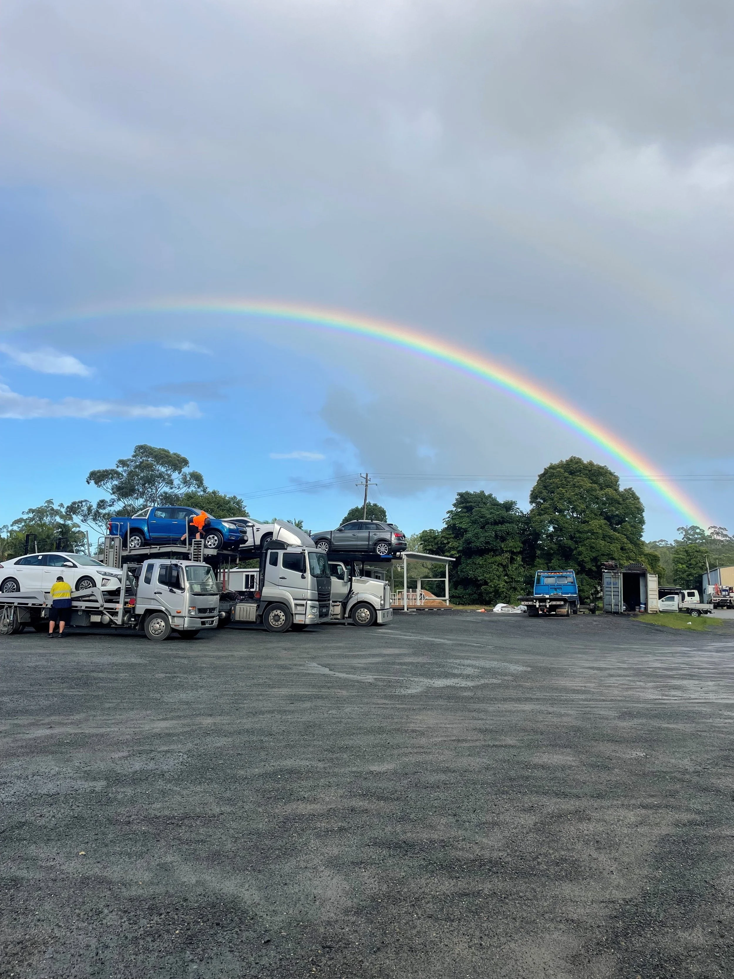 A fleet of cars on a transport truck parked on a gravel lot under a rainbow with partly cloudy skies and trees in the background.