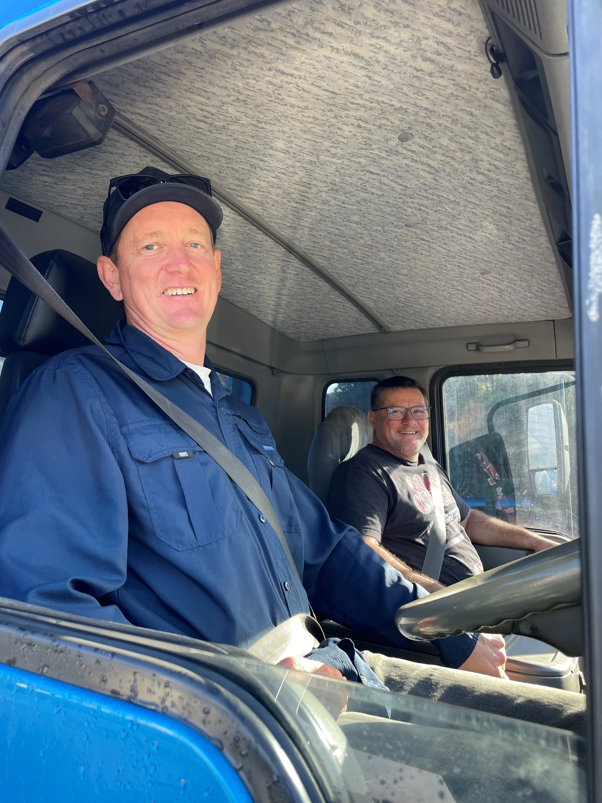 Two men sitting in the front seats of a truck, smiling at the camera. The man on the left is wearing a blue jacket and a black cap, and the man on the right is wearing glasses and a black T-shirt. The truck has a textured ceiling and a blue exterior.
