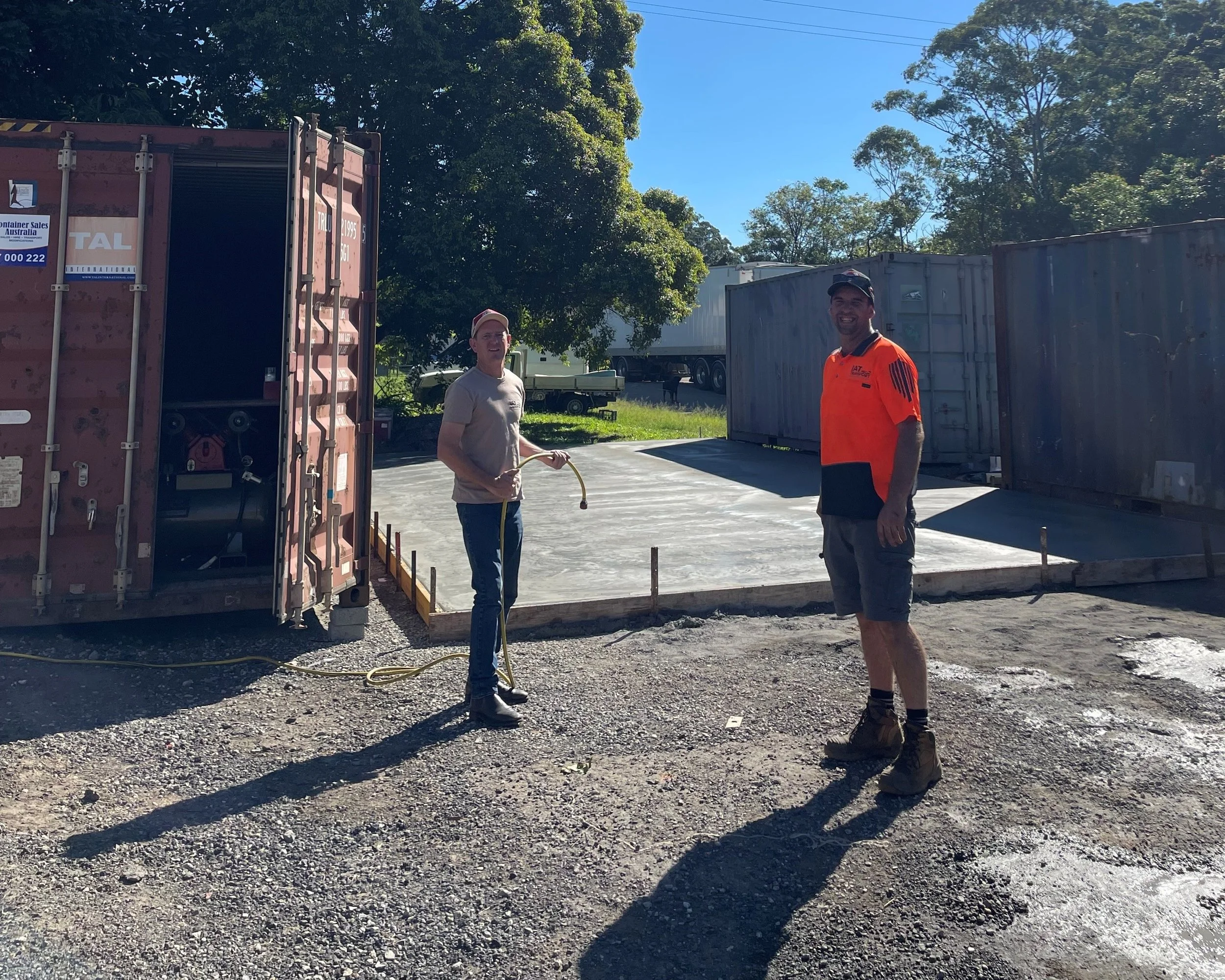 Two men standing on a gravel surface near a concrete slab, construction site with storage containers and trees in the background, one holding a yellow hose, sunny day.