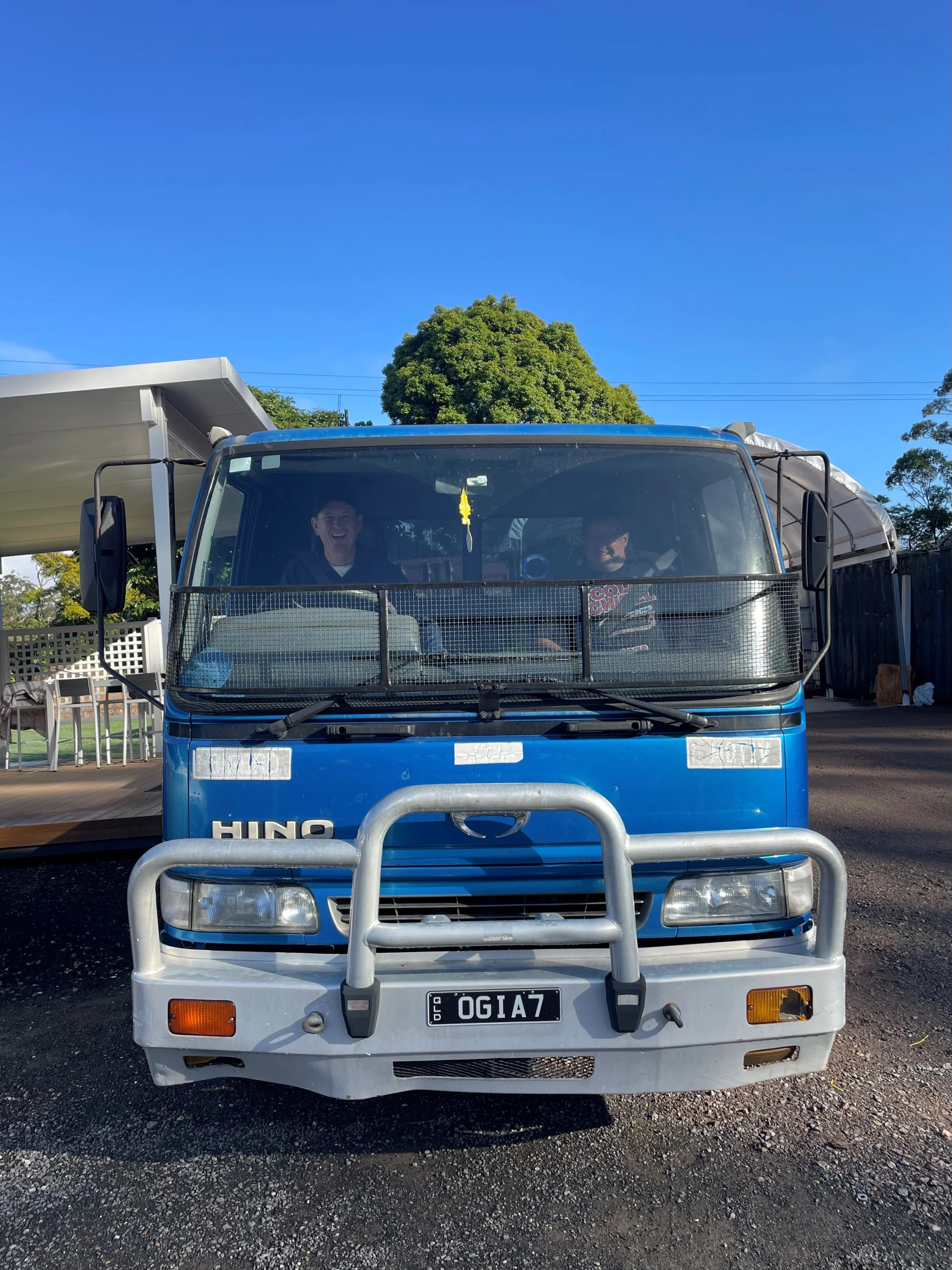 Front view of a blue Hino truck with a silver bull bar and two people inside, smiling. The truck has a black grille, clear headlights, and a Queensland license plate reading 01IA7. The background shows a clear blue sky, green trees, and outdoor furniture and structures.