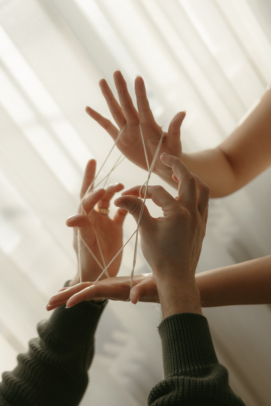 Close-up of multiple hands intertwined with yarn, creating a complex pattern against a light background.