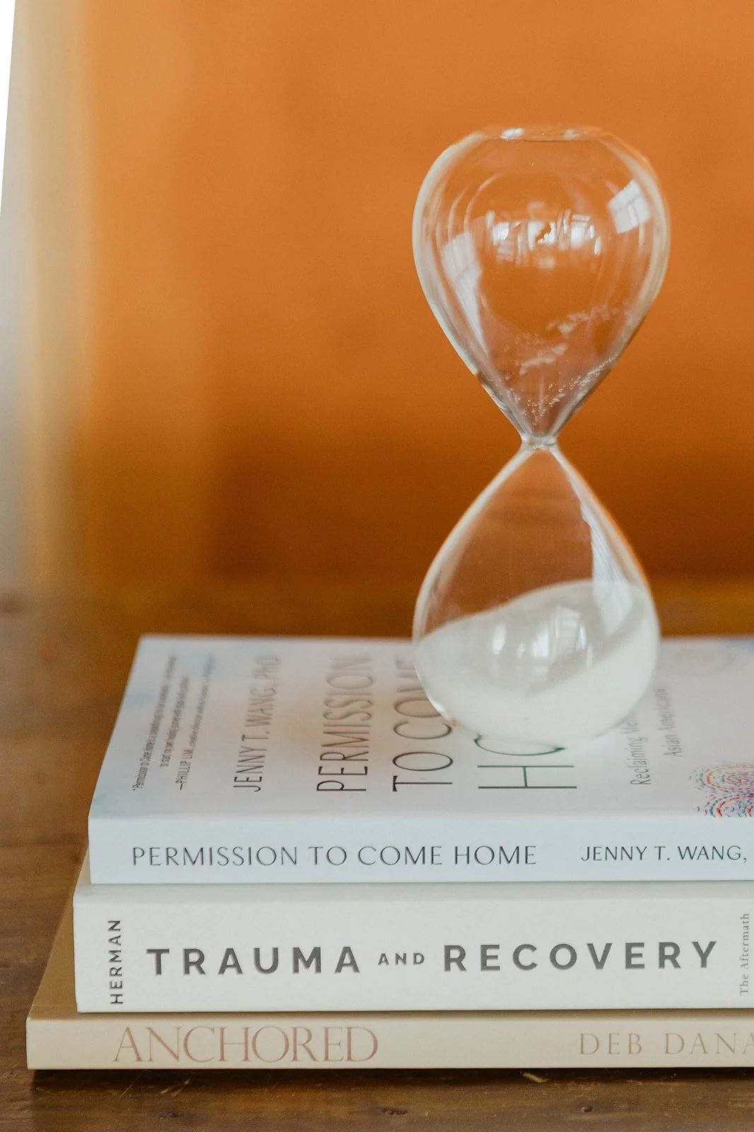 An hourglass resting on top of a stack of three books with a plain orange wall in the background.