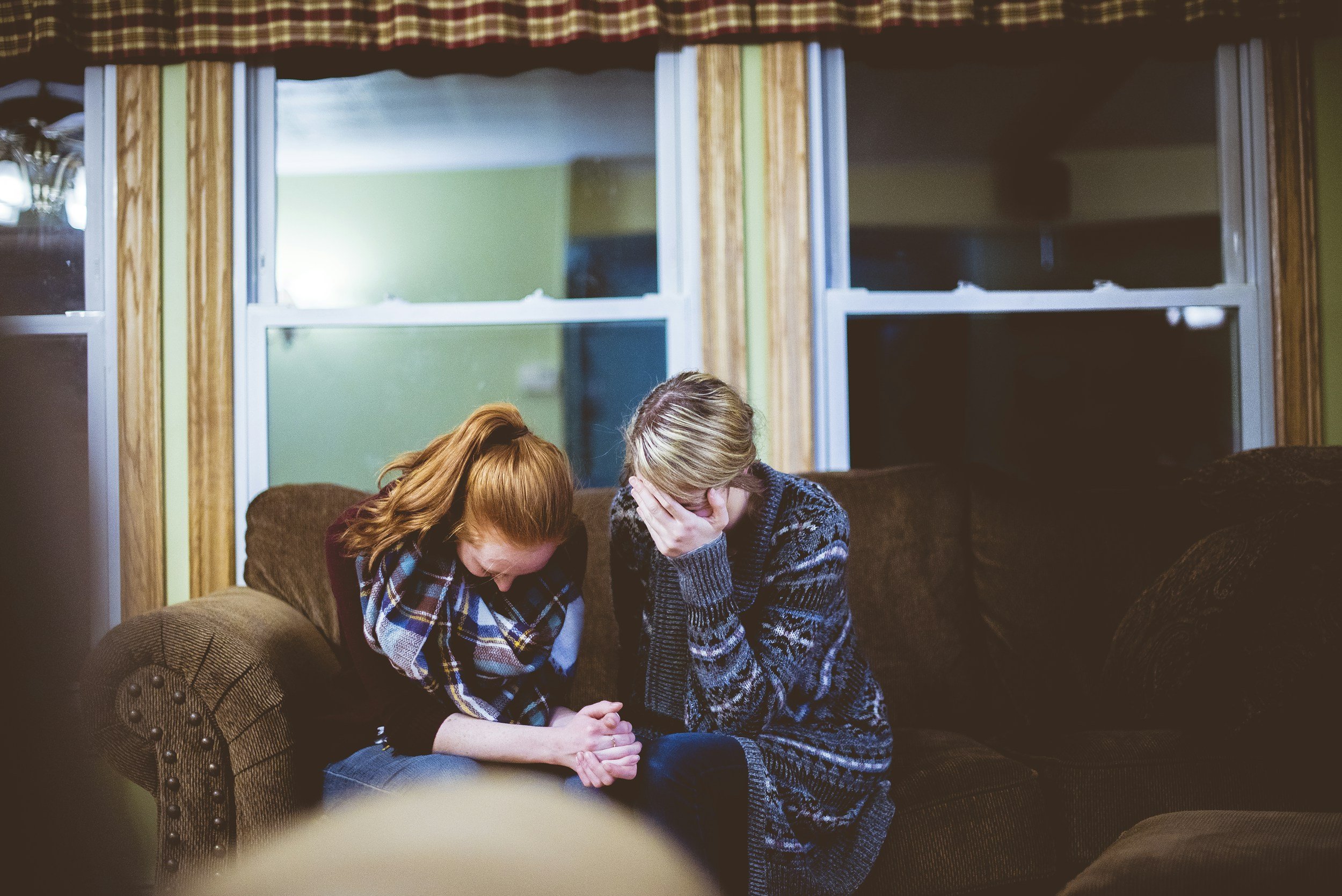 Two women sitting on a brown couch with their heads bowed and hands clasped together in prayer, inside a living room with green walls, wooden window frames, and a window with curtains.