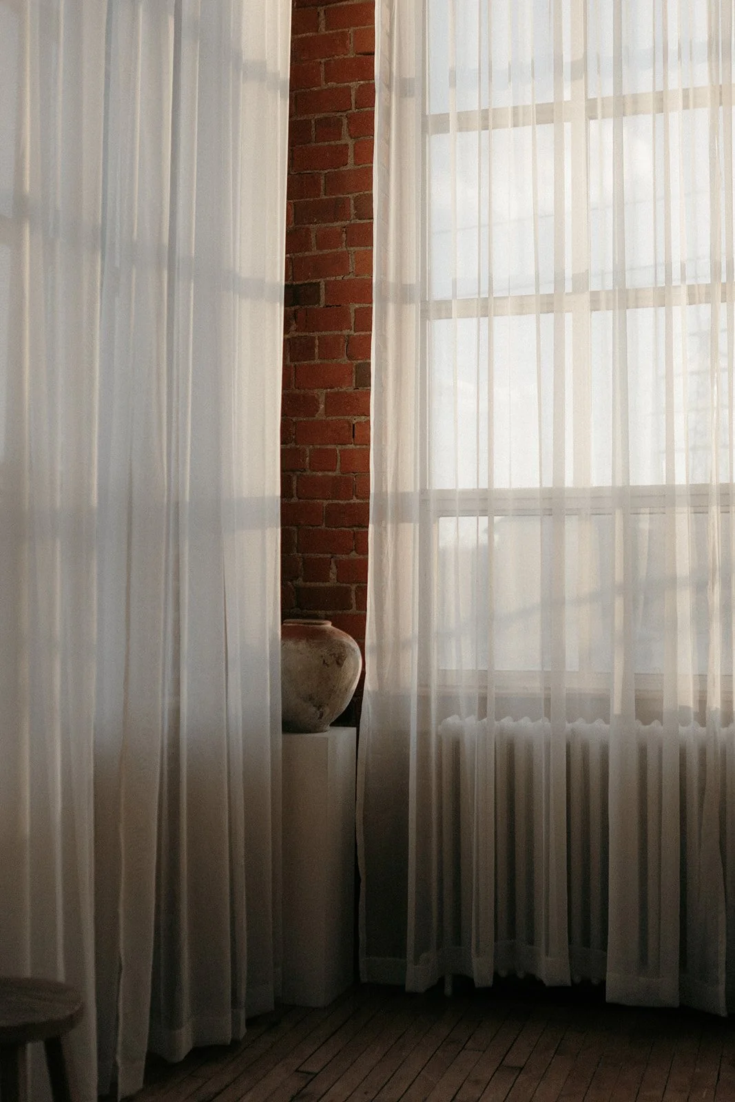 Interior view of a room with sheer white curtains, a brick wall, a large window, and a ceramic vase on a pedestal.