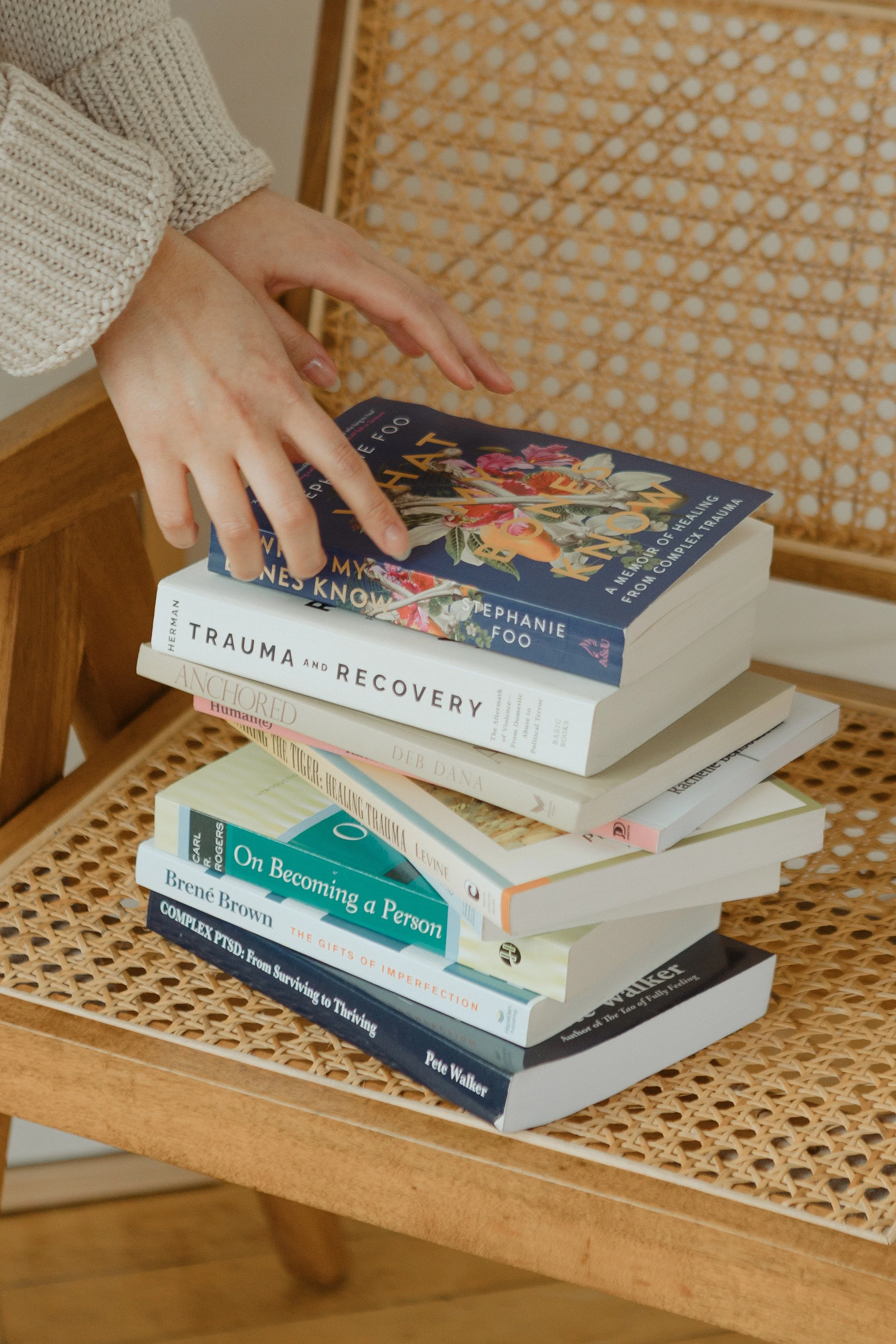 A stack of books on a wooden table with a person flipping through a book. The books have titles related to trauma, healing, personality, and mental health.