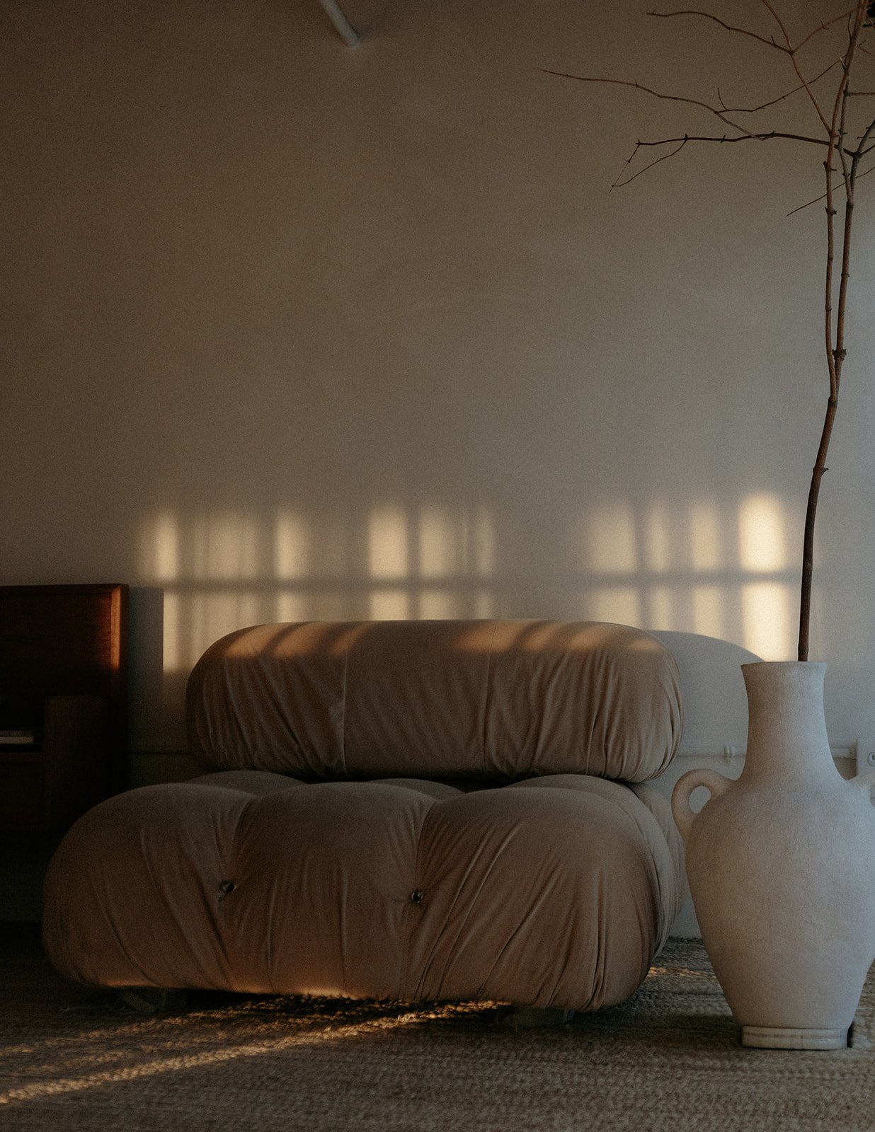 A beige tufted armchair in front of a wall with sunlight and shadows cast through window panes, beside a ceramic vase with a tall, leafless branch.