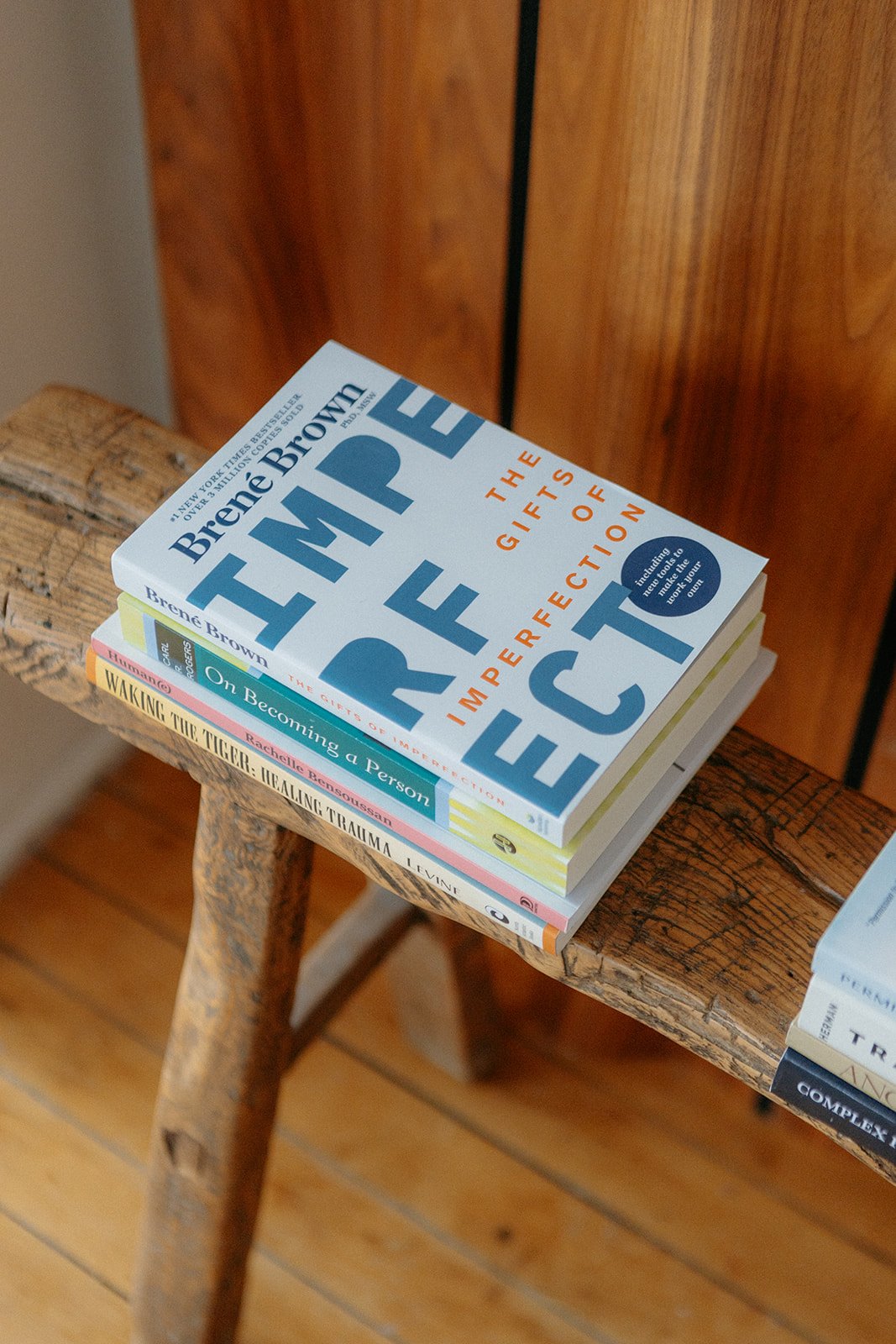 Stack of four books on a rustic wooden chair. The top book is titled 'The Gifts of Imperfection' by Brené Brown. The second is 'On Becoming a Person' by Carl Rogers, the third is 'Waking the Tiger' by Peter Levine, and the fourth appears to be related to trauma healing.