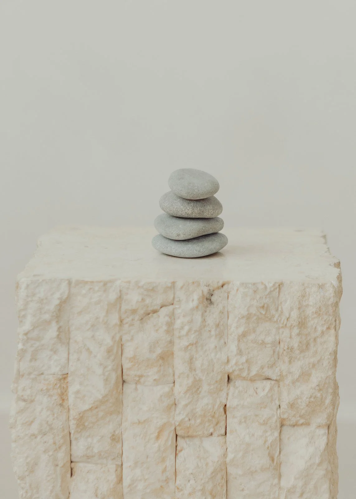 Five smooth gray stones stacked on top of a white textured block against a plain light background.
