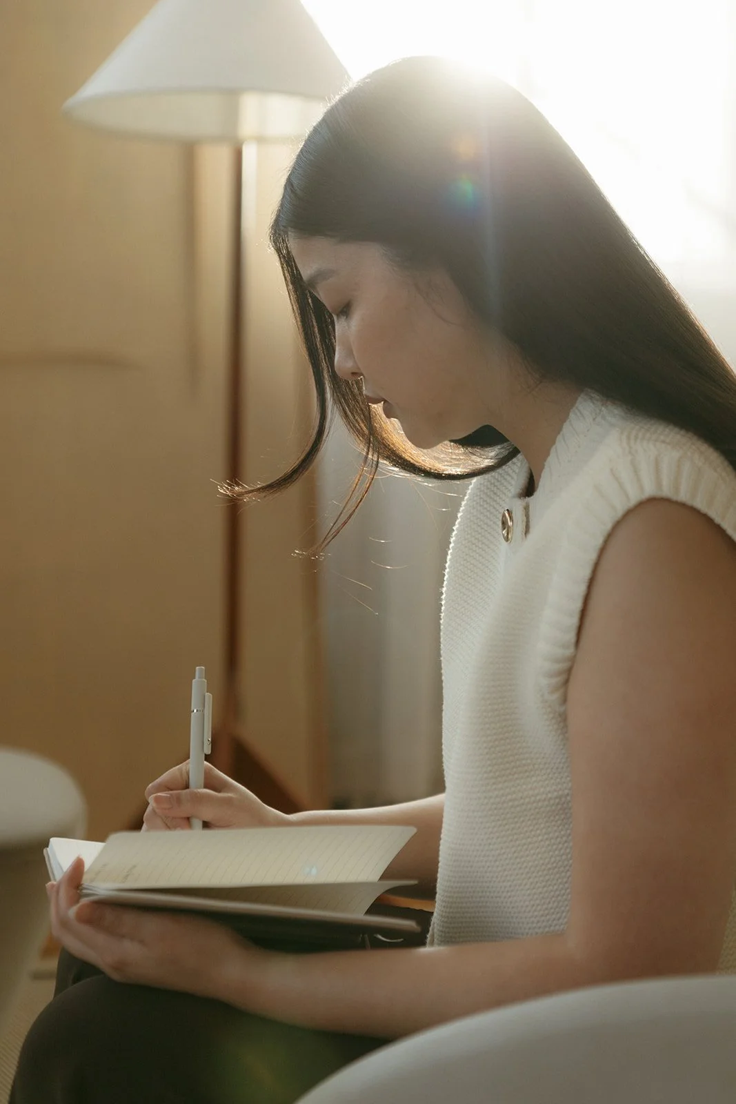 A woman with long dark hair, wearing a sleeveless white top, is sitting and writing in a notebook with a pen in her right hand. Sunlight streams behind her, creating a bright, warm atmosphere.