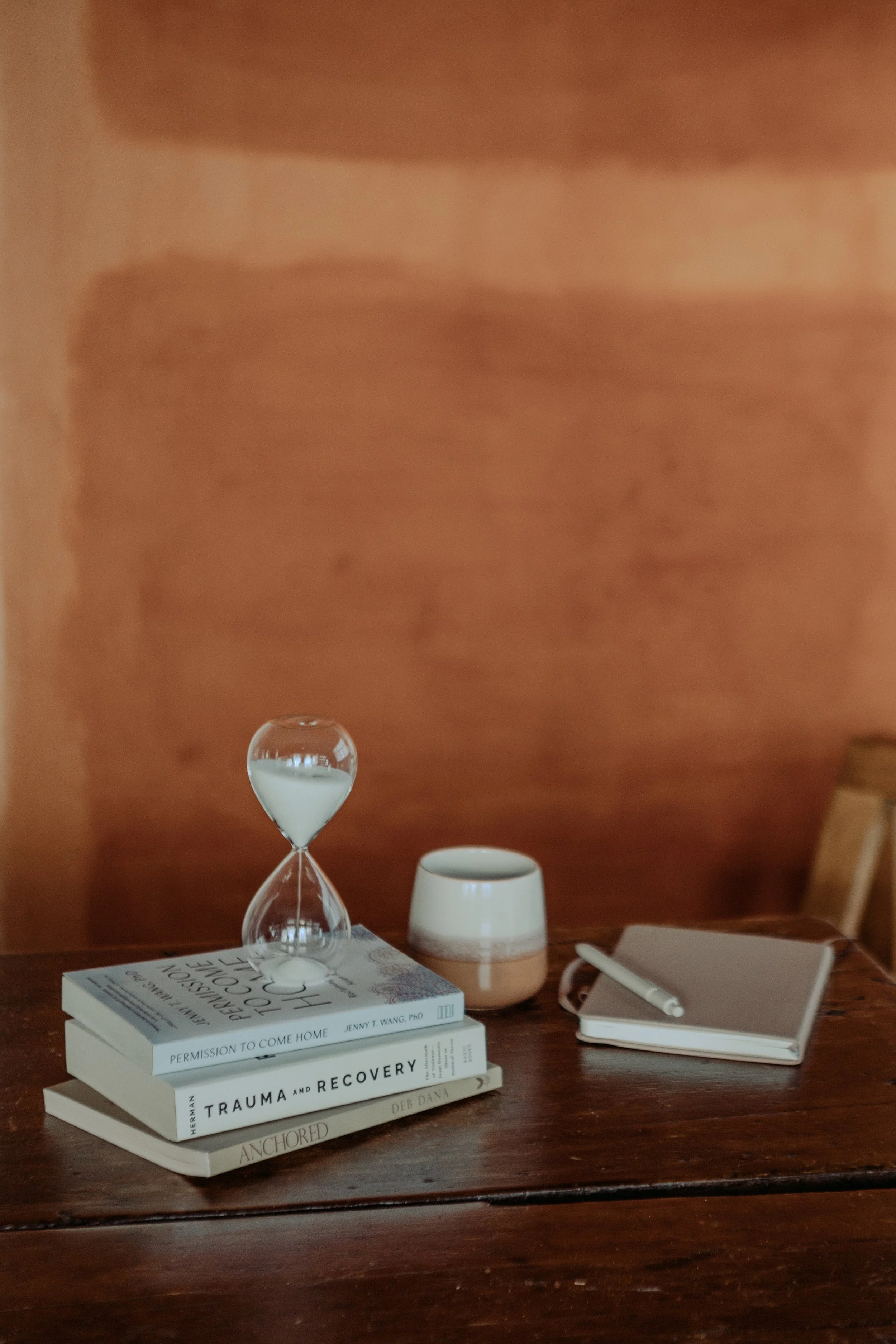 A wooden table with a stack of books, a white ceramic cup, a glass hourglass, a notebook, and a pen, with a blurred wooden wall in the background.