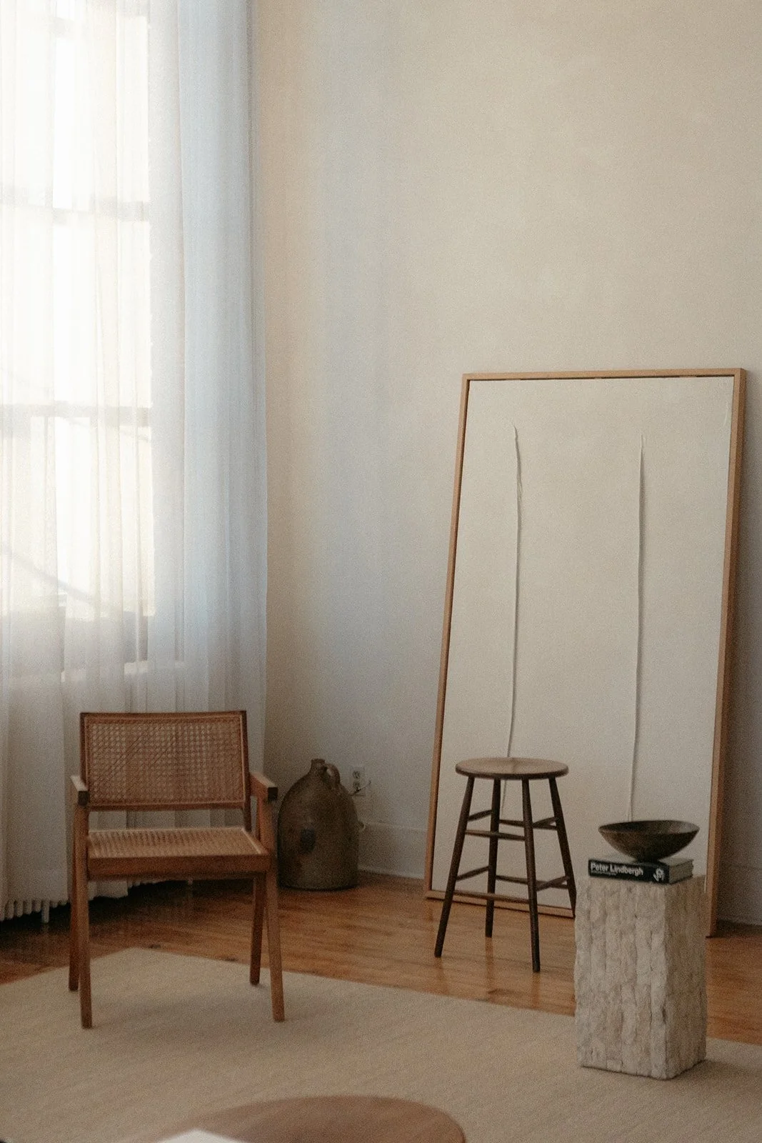 Minimalist interior with a wooden chair, a large framed mirror, a small stool, a stone pedestal with a book and bowl, a ceramic vase, and sheer curtains at a window.
