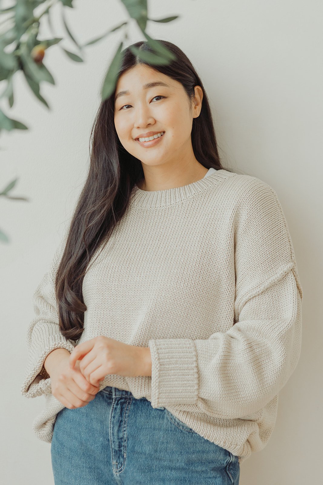 A young woman with long dark hair smiling, wearing a cream-colored sweater and blue jeans, standing against a light-colored wall with some green leaves partially in the frame.