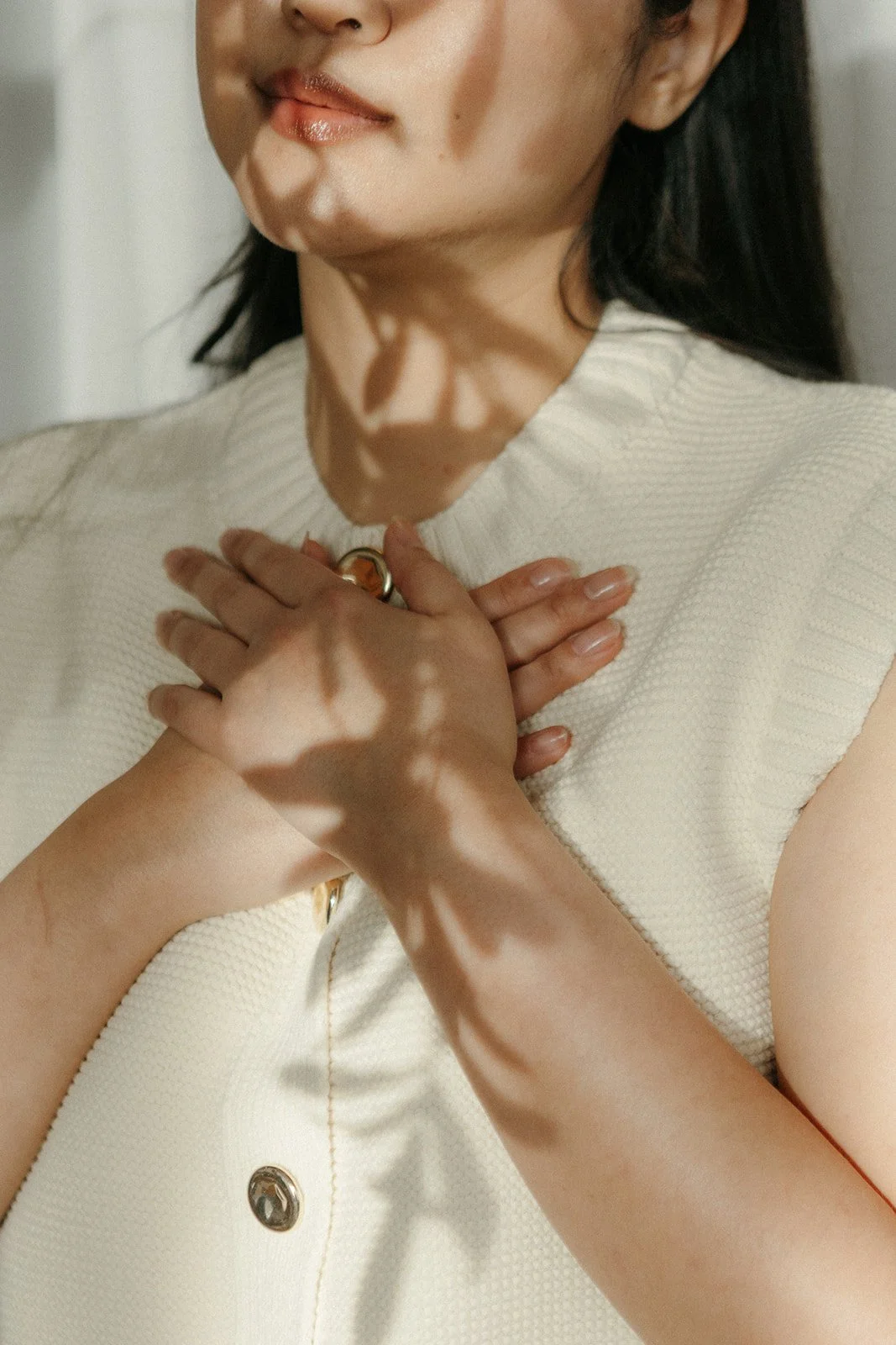 A woman with light skin and dark hair wearing a sleeveless white top, holding her hand over her chest. Sunlight creates shadows of her hand and face.