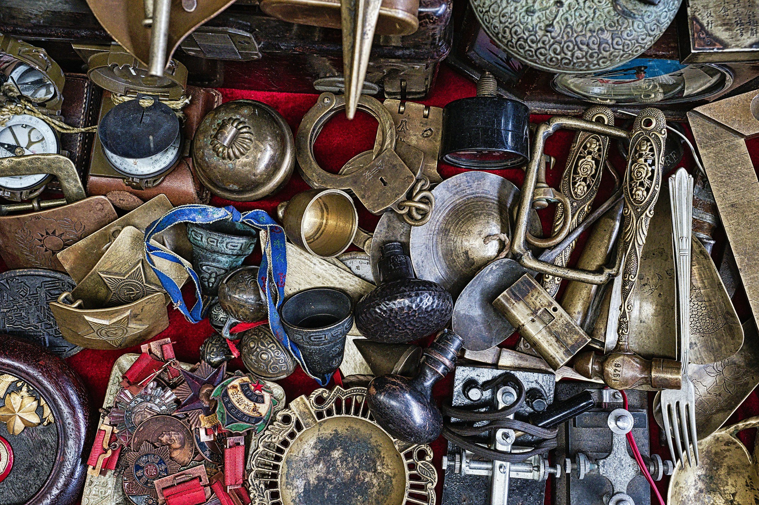 A collection of vintage and antique items including pocket watches, medals, handcuffs, brass objects, cutlery, and metal tools arranged on a red surface.