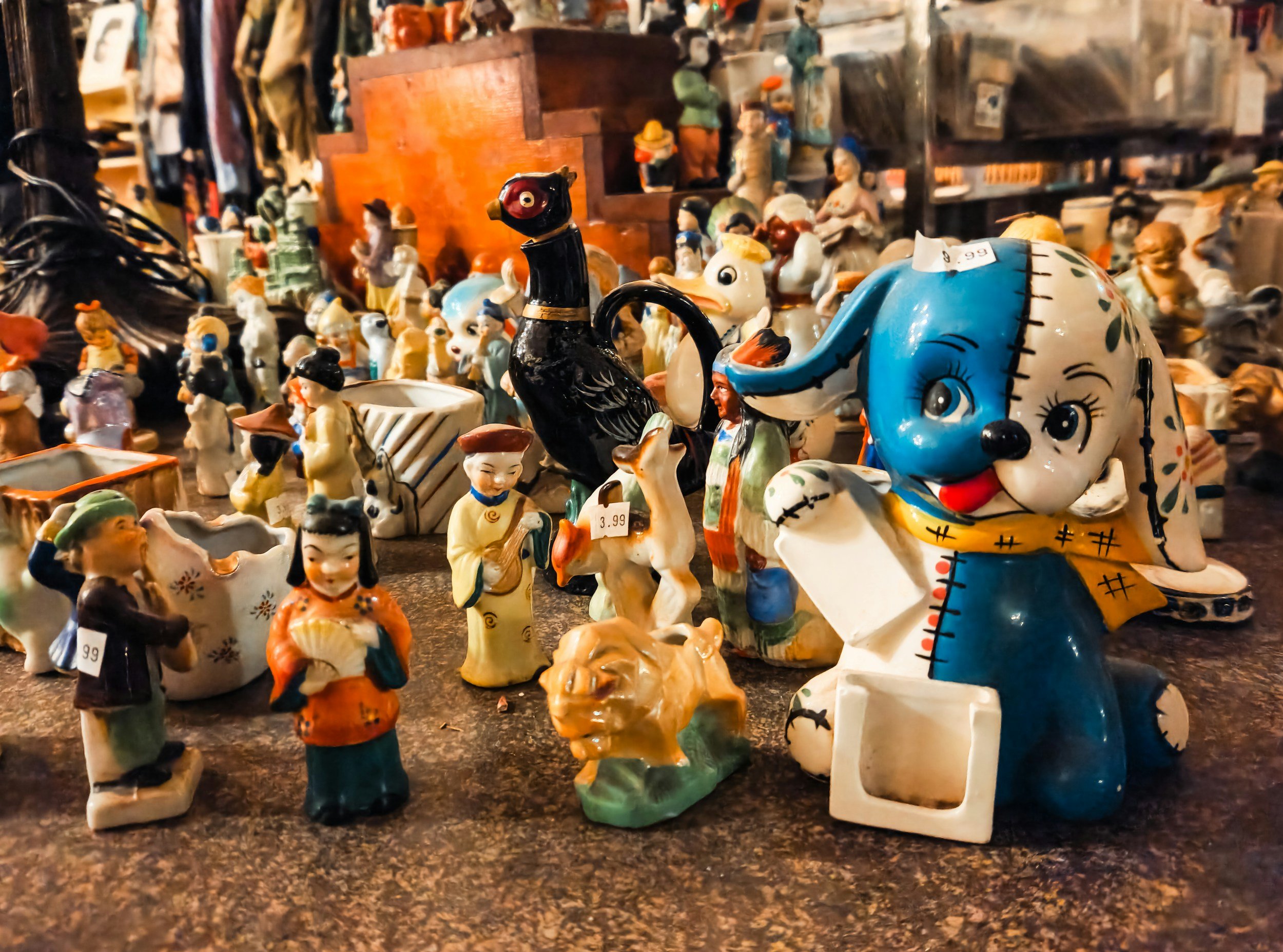 Collection of small, colorful ceramic figurines including a dog, a lion, a woman, and various other characters, displayed on a dark surface in a shop.