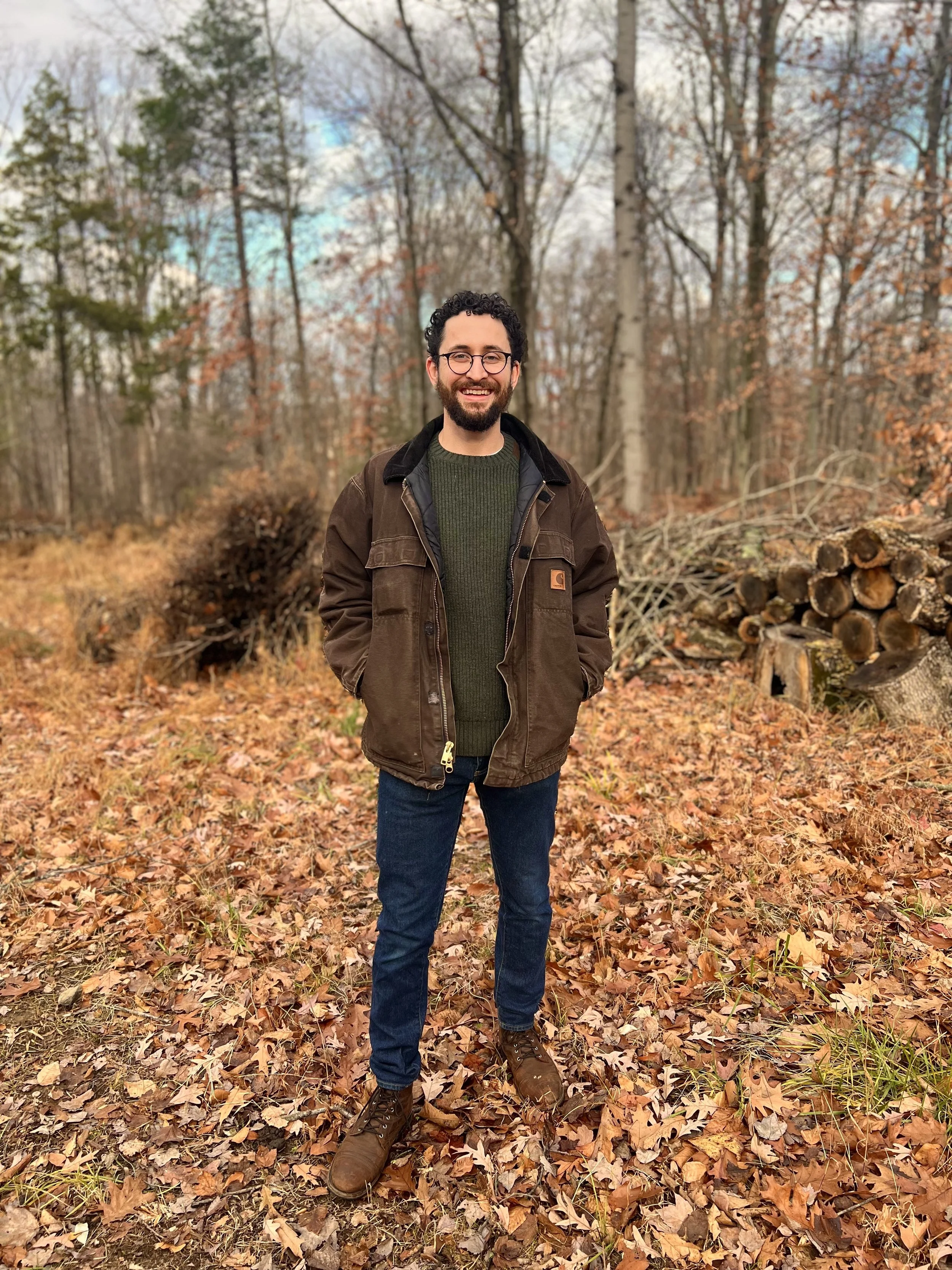 A man smiling outdoors in a wooded area with fallen leaves, wearing a brown jacket, green sweater, jeans, and boots.