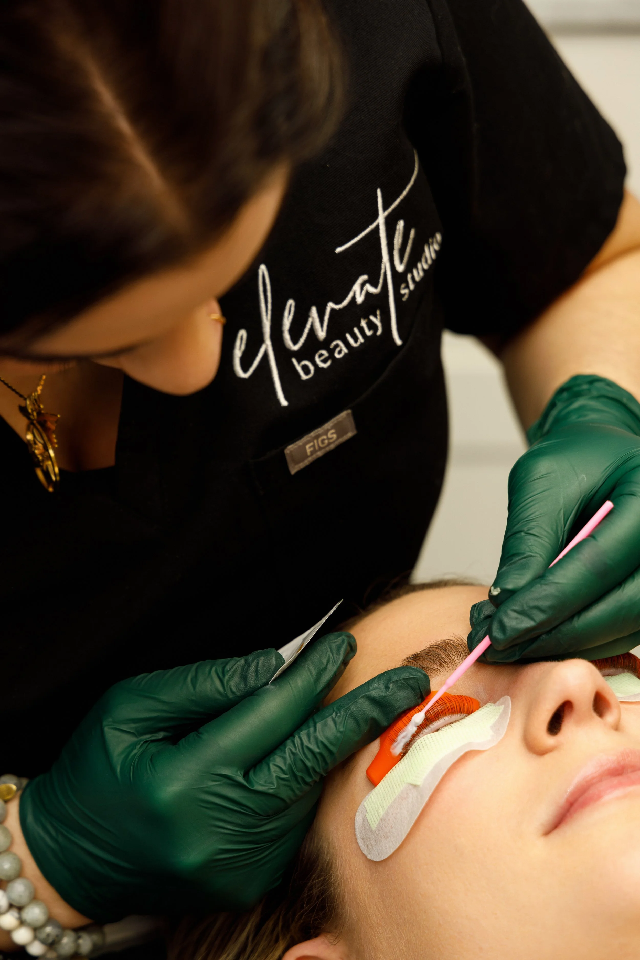 Lash Lift solution being applied to a female model by a female esthetician wearing a scrub top that has the logo of elevate beauty studio embroidered on it.