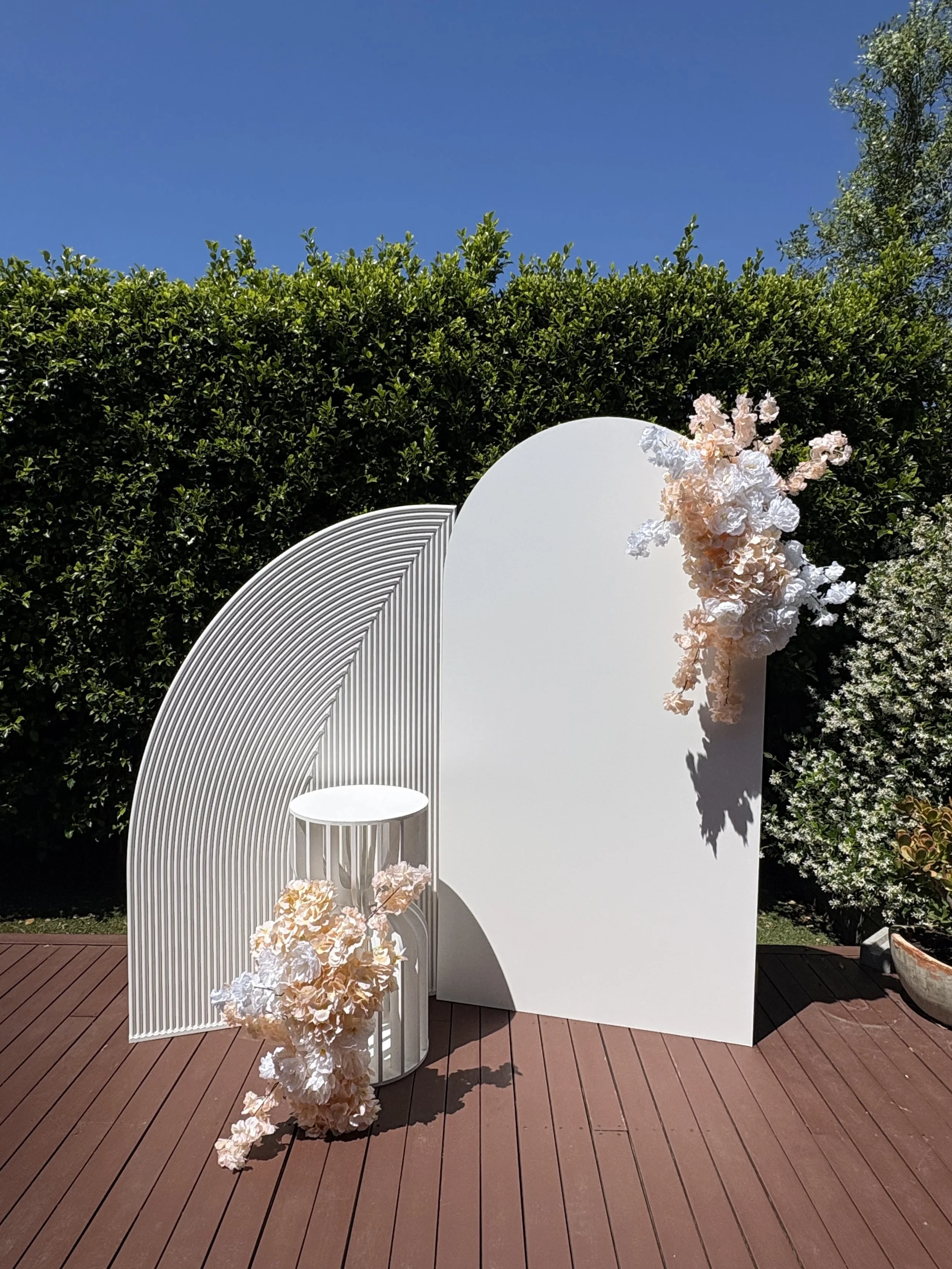 A decorative outdoor setup on a wooden deck featuring white curved panels, a white stool with floral arrangements of soft pink and white flowers, and a lush green hedge under a clear blue sky.