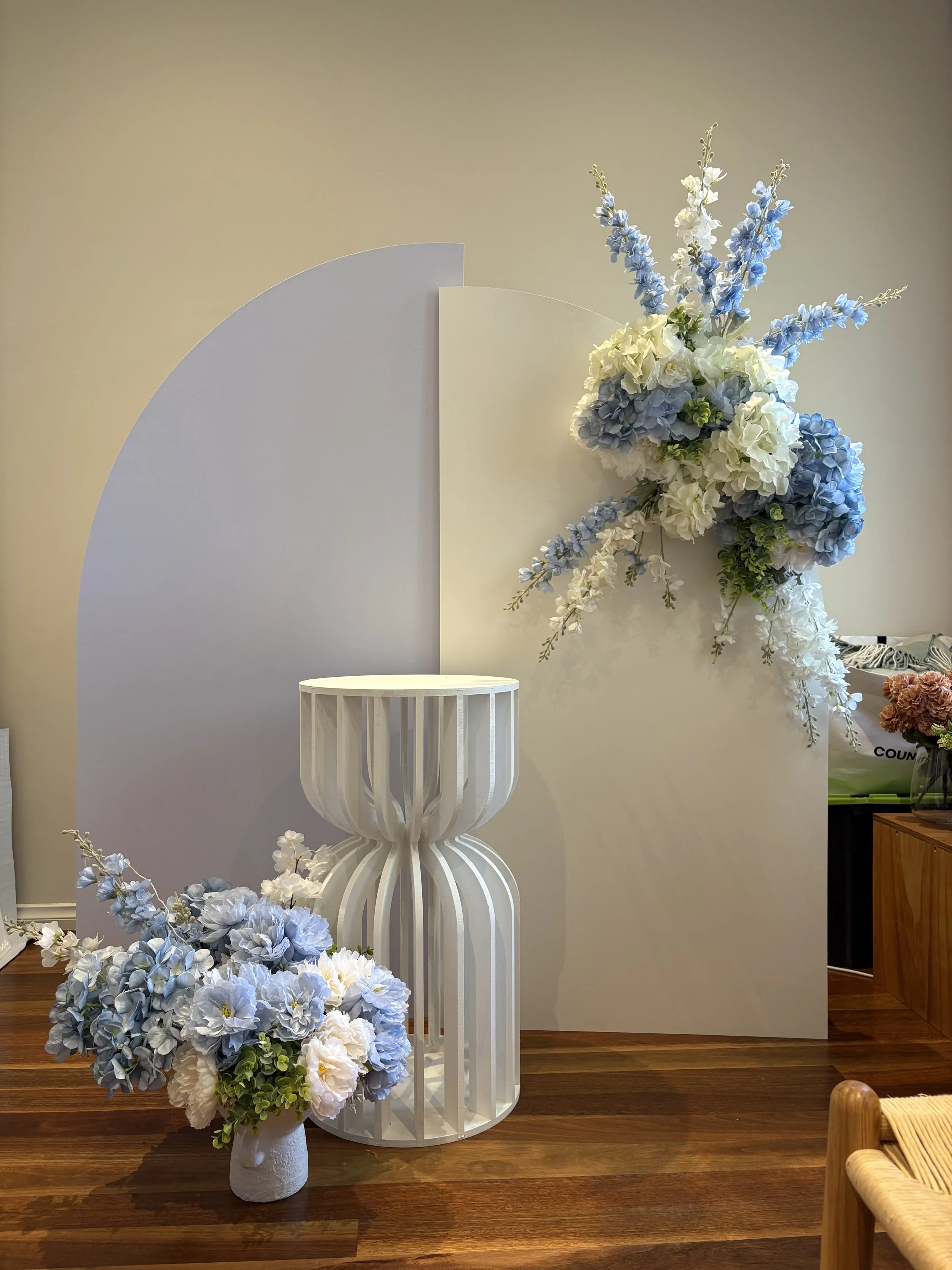 A floral arrangement with white and blue flowers on a white geometric stand, and a smaller bouquet of similar flowers in a white vase on a wooden floor, with a backdrop partially painted in white and lavender.