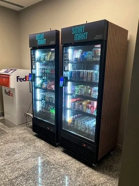 Two 24-hour vending machines stocked with snacks and beverages, located in a corner of a room with wooden flooring and beige walls.