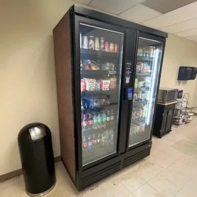 Two vending machines labeled '24/7 Smart Vending' filled with snacks and beverages, located against a beige wall on a light wood floor.