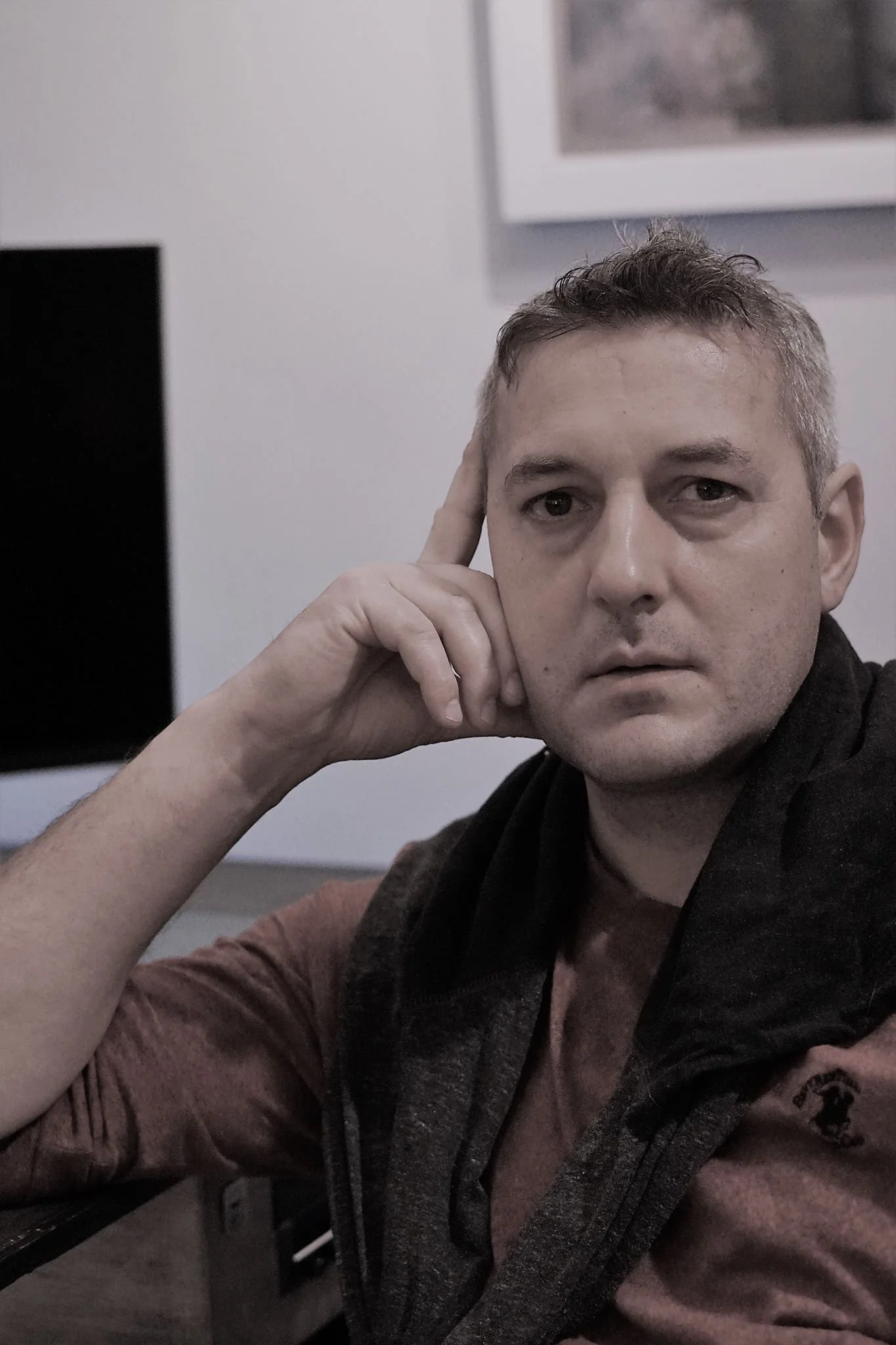 A man sitting at a desk, resting his head on his hand and looking at the camera with a serious expression.