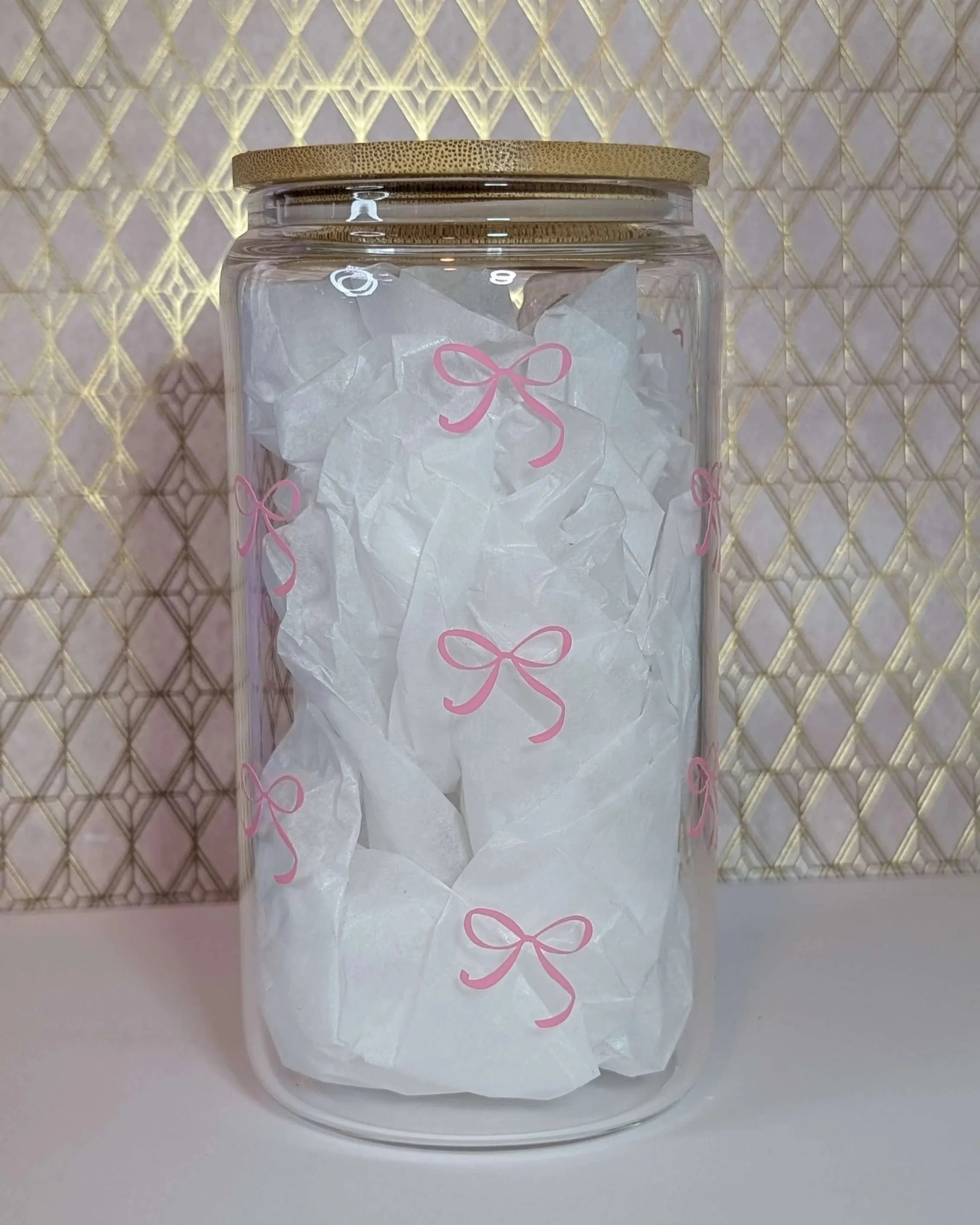 A glass jar with a gold lid filled with crumpled tissue paper decorated with pink bow prints, placed against a patterned gold and pink background.