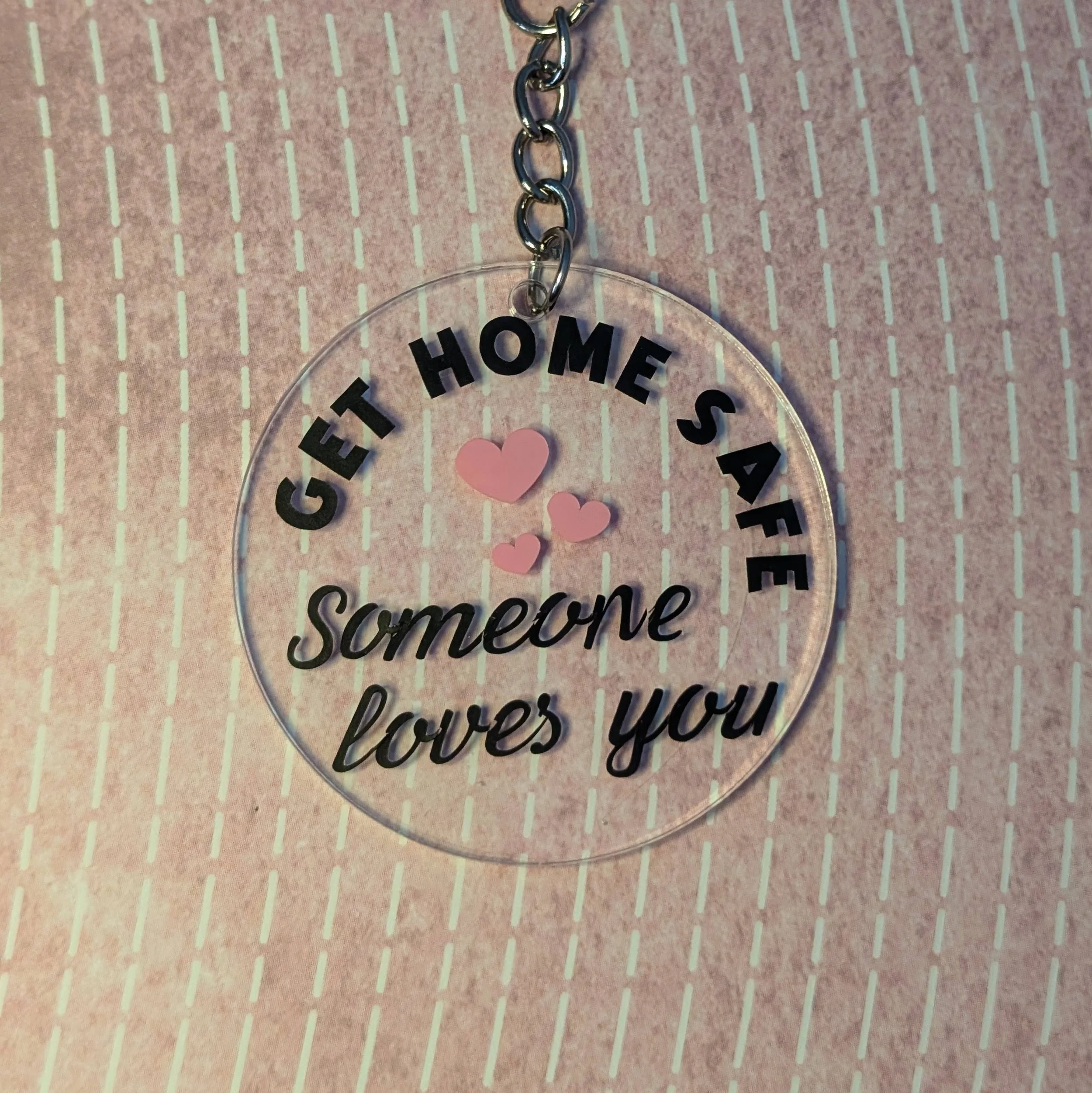 A clear circular keychain with black text that reads 'Get Home Safe, Someone loves you', decorated with three pink hearts, hangs on a silver chain against a pink background with white dashed lines.