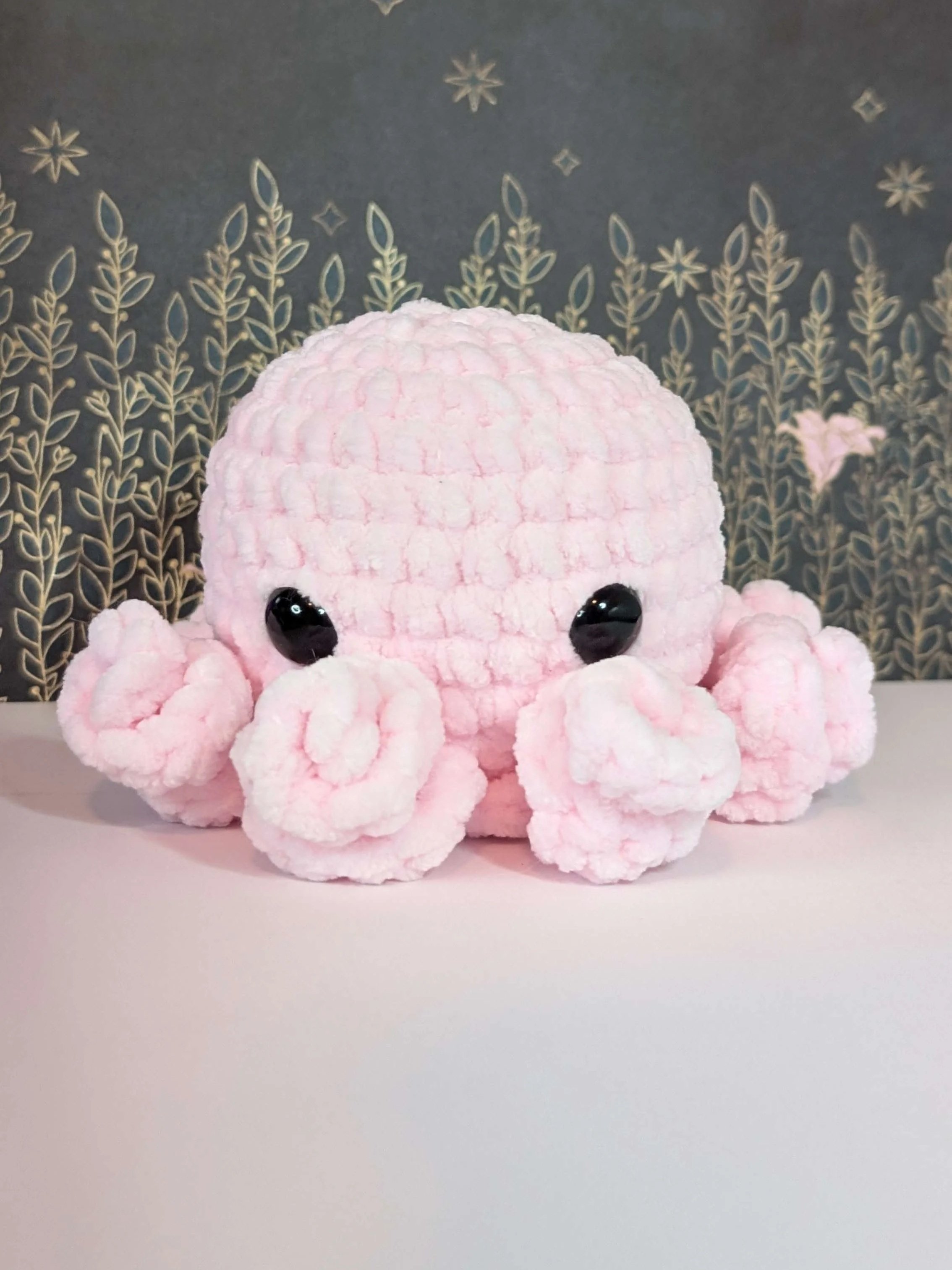 A pink plush octopus toy with large black eyes on a white surface, with a decorative dark background featuring a floral and star pattern.