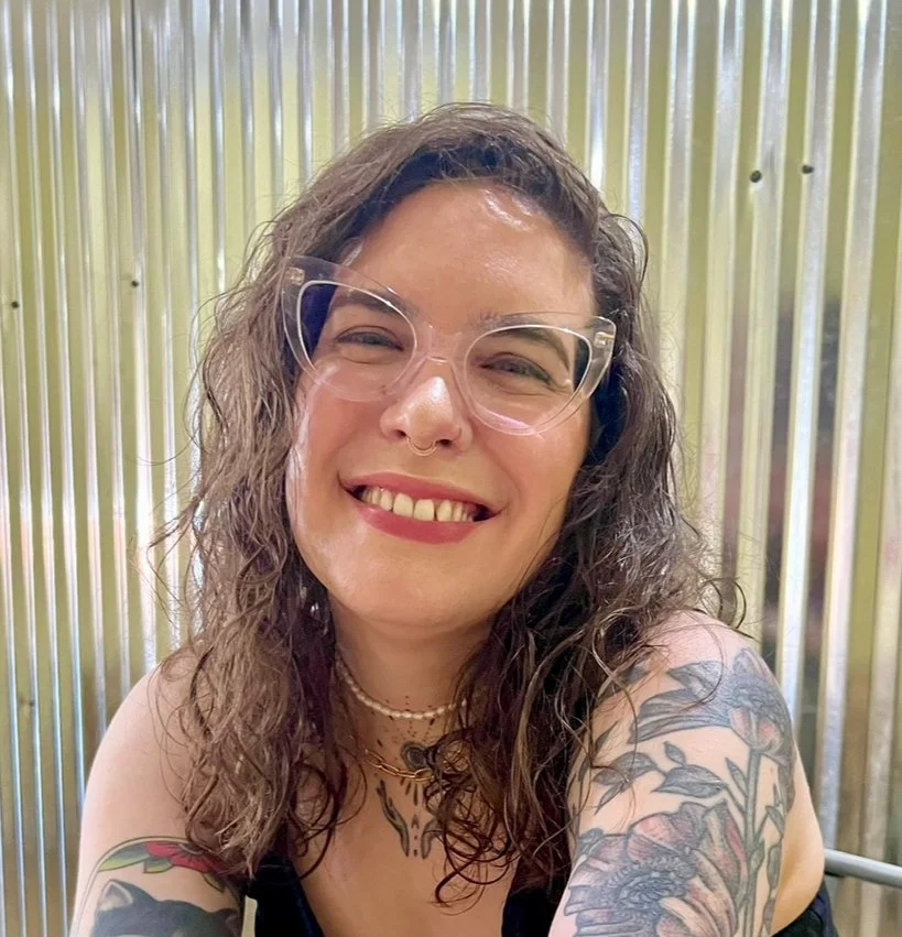 Close-up of a smiling woman with wavy hair, wearing clear glasses, a nose ring, layered necklaces, and showing tattoos on her shoulders and chest, standing in front of a metallic, corrugated background.