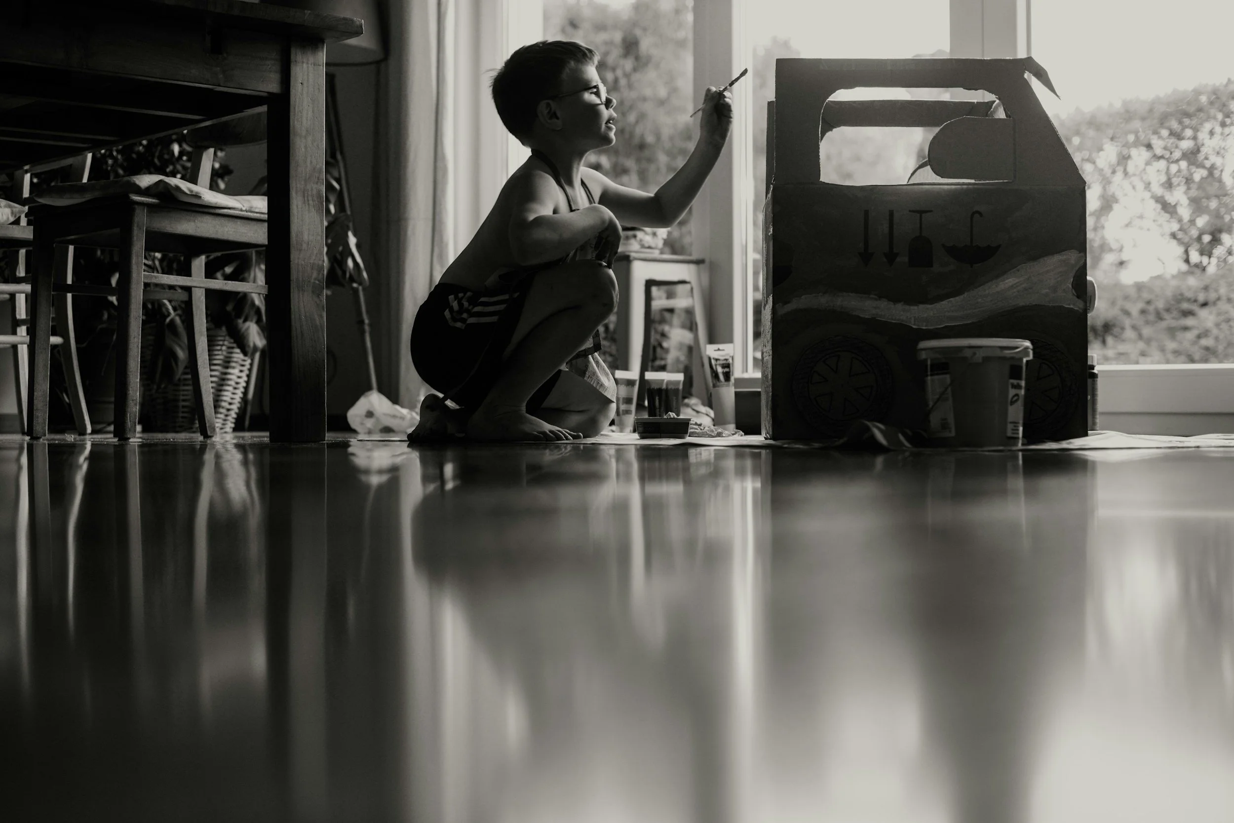 A child crouching on a hardwood floor, drawing on a cardboard box near a large window, with a table and chairs in the background.