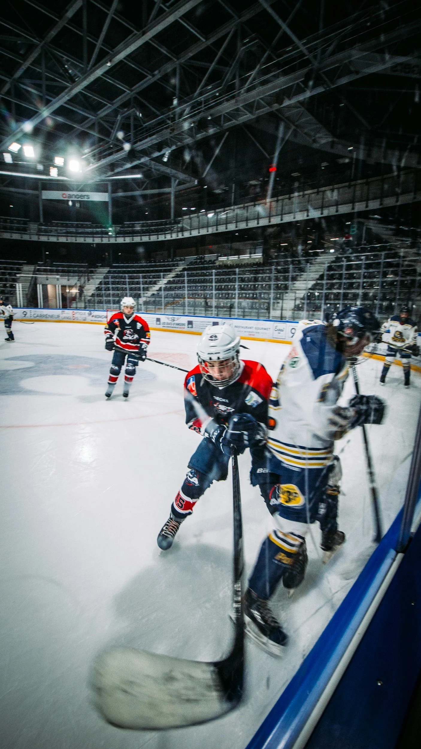 Hockey players in a game on an ice rink inside an arena, with some players near the boards and others in the background, and spectators' seats visible in the upper stands.