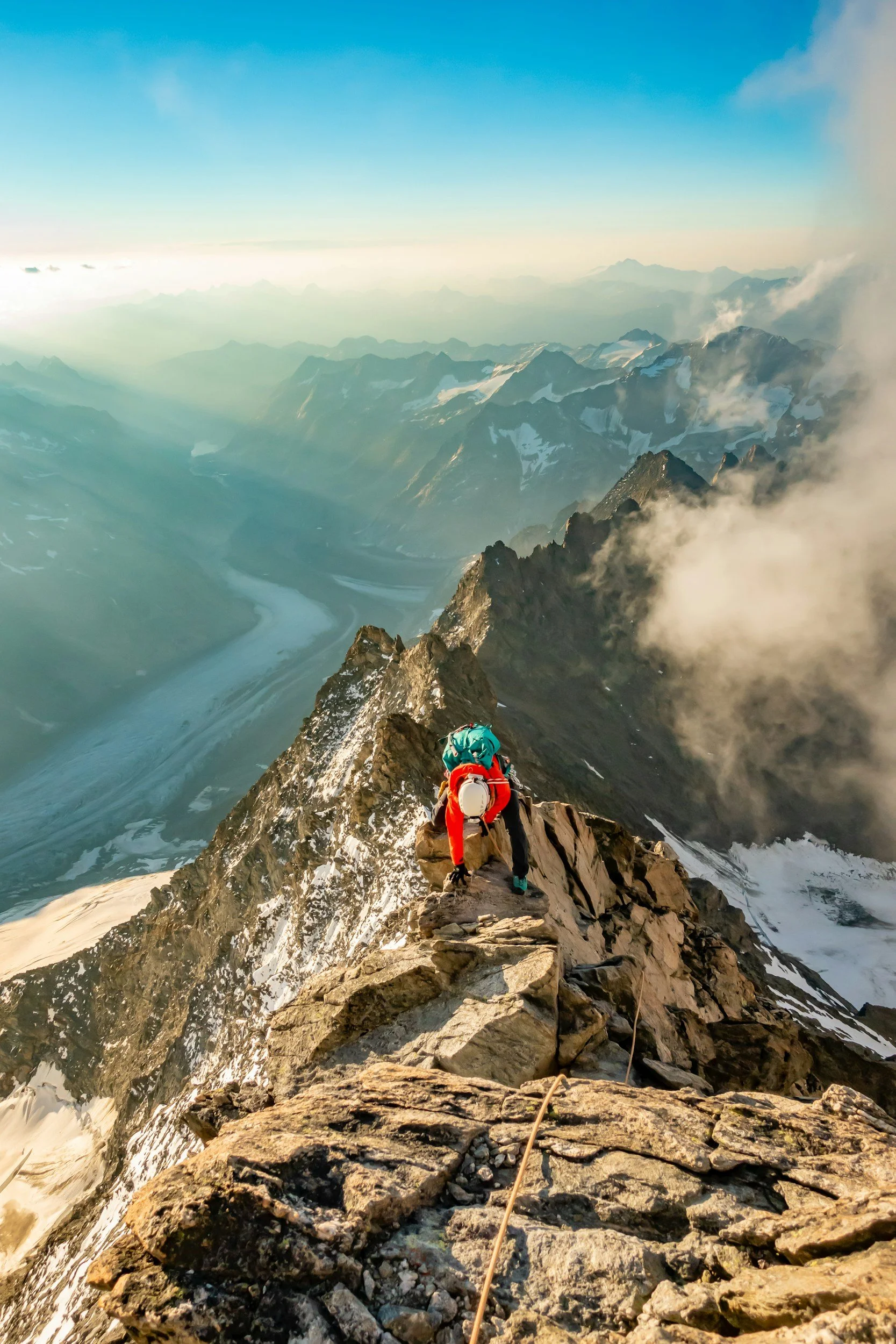 A person climbing a rocky mountain ridge with snow-capped peaks in the background, clouds, and a river far below.