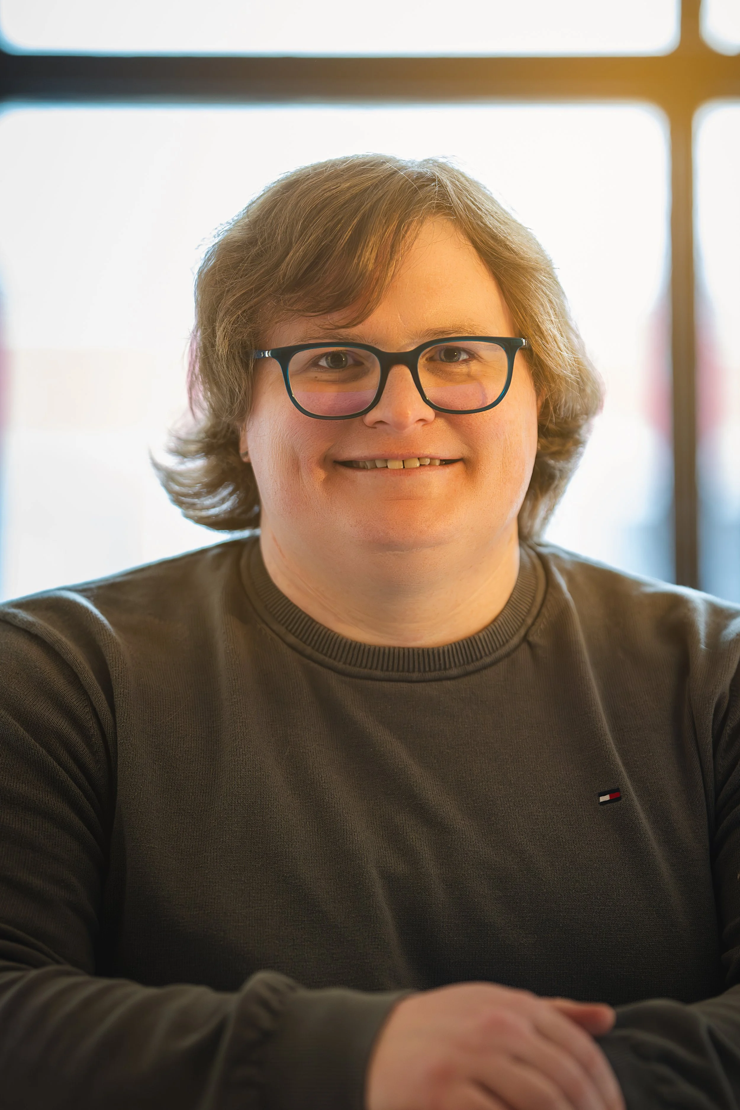 A person with medium-length, wavy hair, glasses, and a dark-colored sweater, smiling and sitting with folded arms in front of a window with sunlight streaming through.