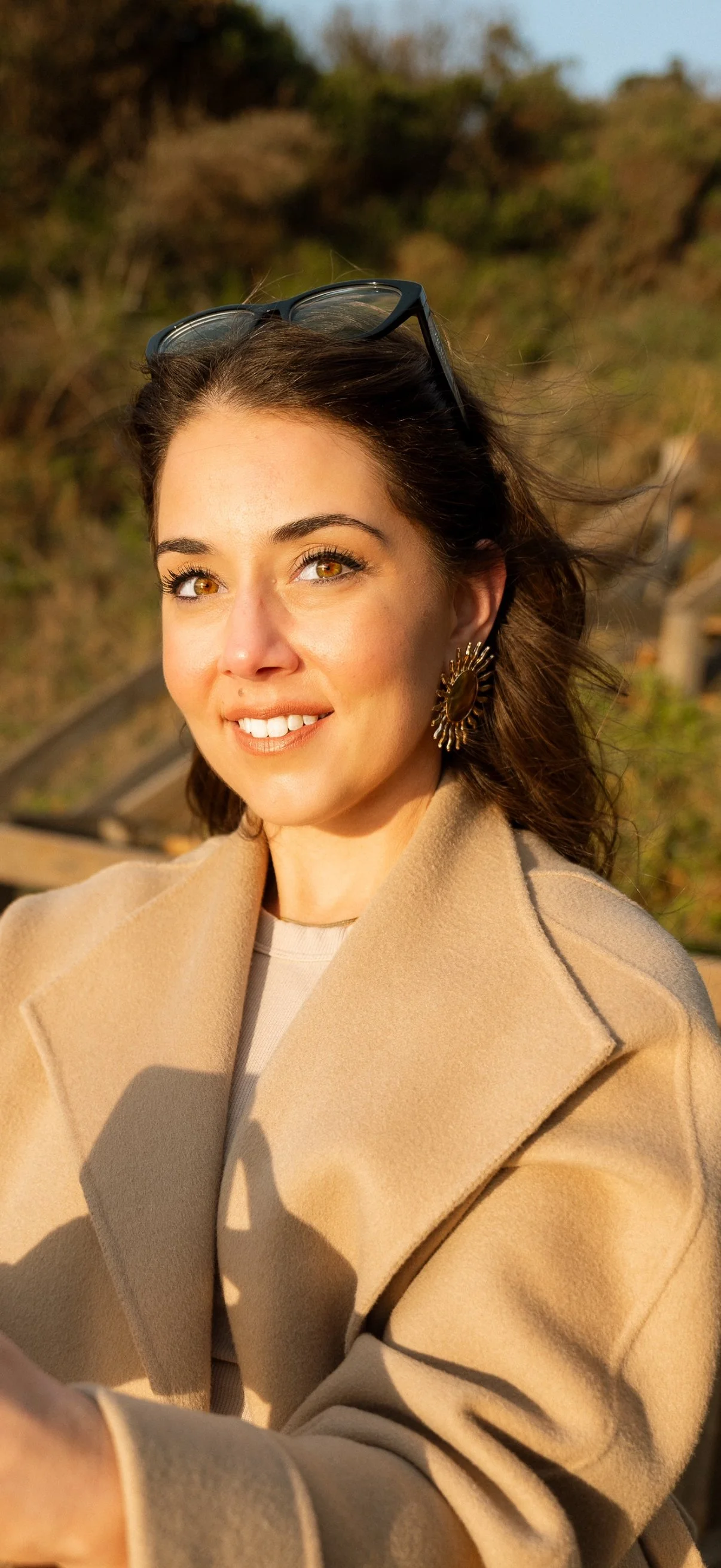 Close-up of a smiling woman with brown hair, wearing large sunflower-shaped earrings, beige coat, and sunglasses on her head, outdoors during sunset.