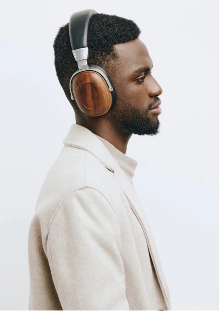 Profile of a man with dark skin wearing large over-ear headphones with wooden ear cups and a light-colored jacket, facing to the right against a plain light background.