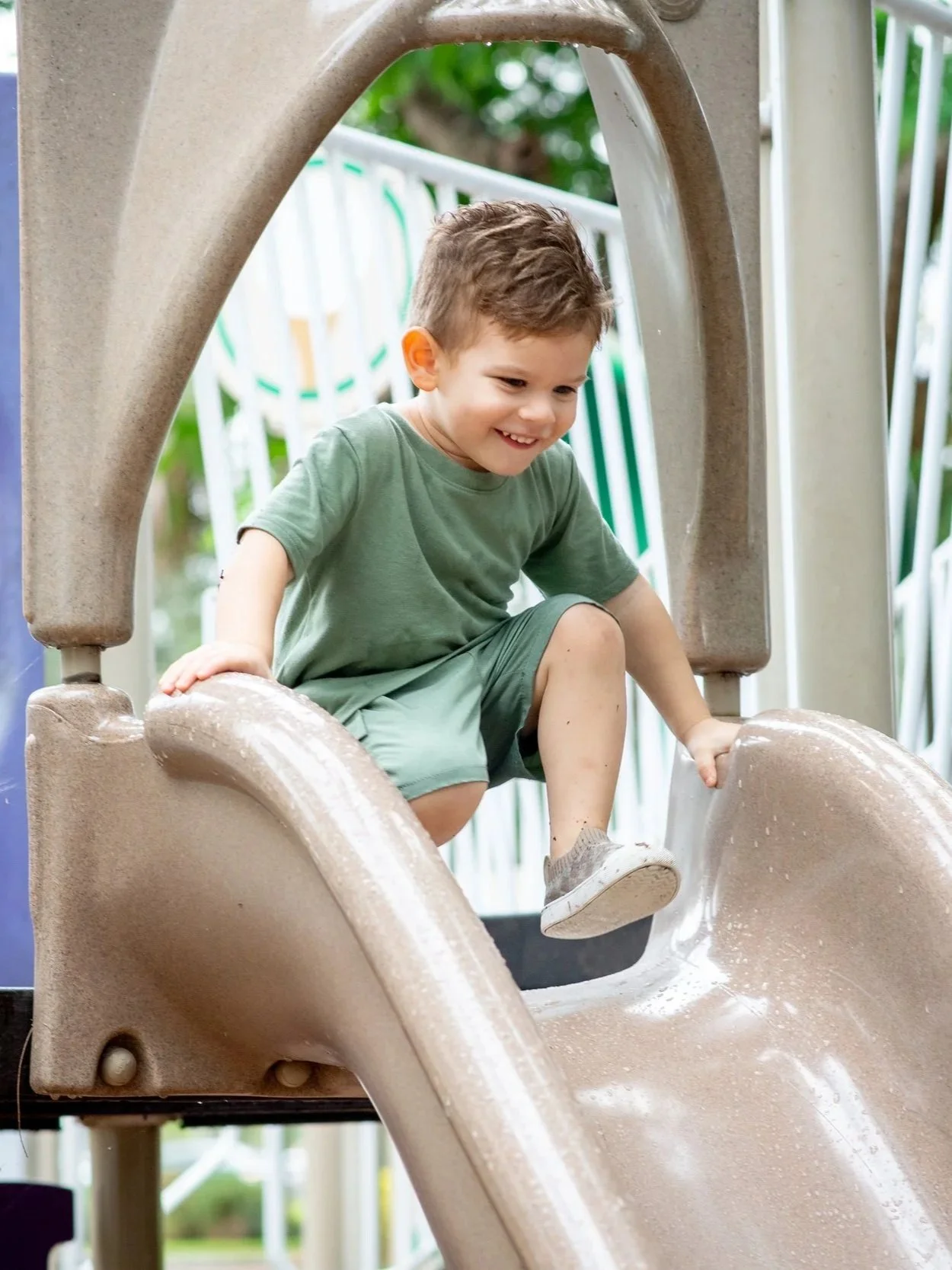 A young boy is sliding down a beige playground slide, smiling and wearing a green t-shirt and shorts.