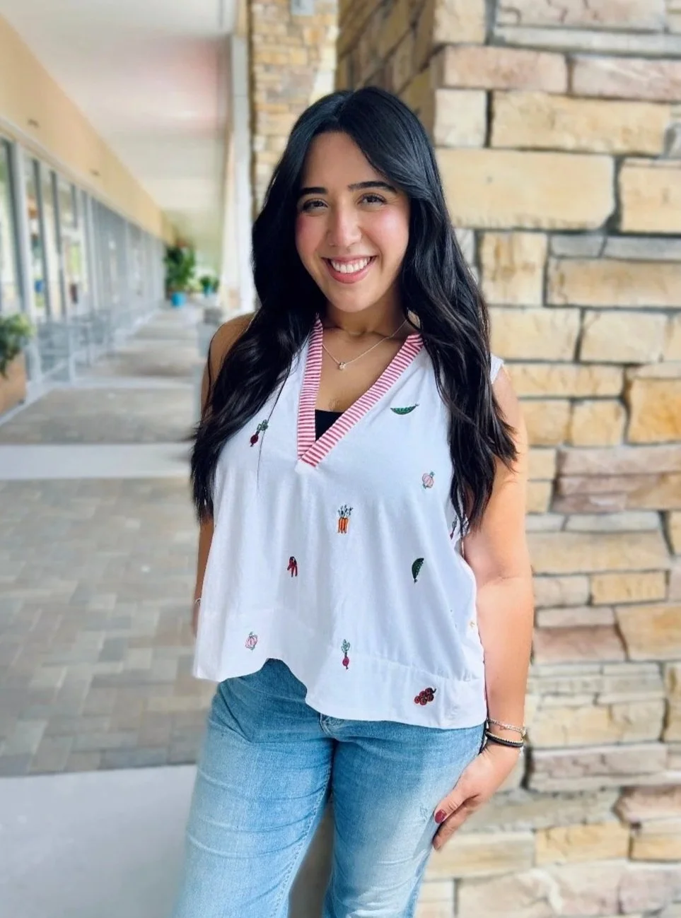 A young woman with long black hair smiling and standing near a brick wall outside a building with large windows and potted plants.
