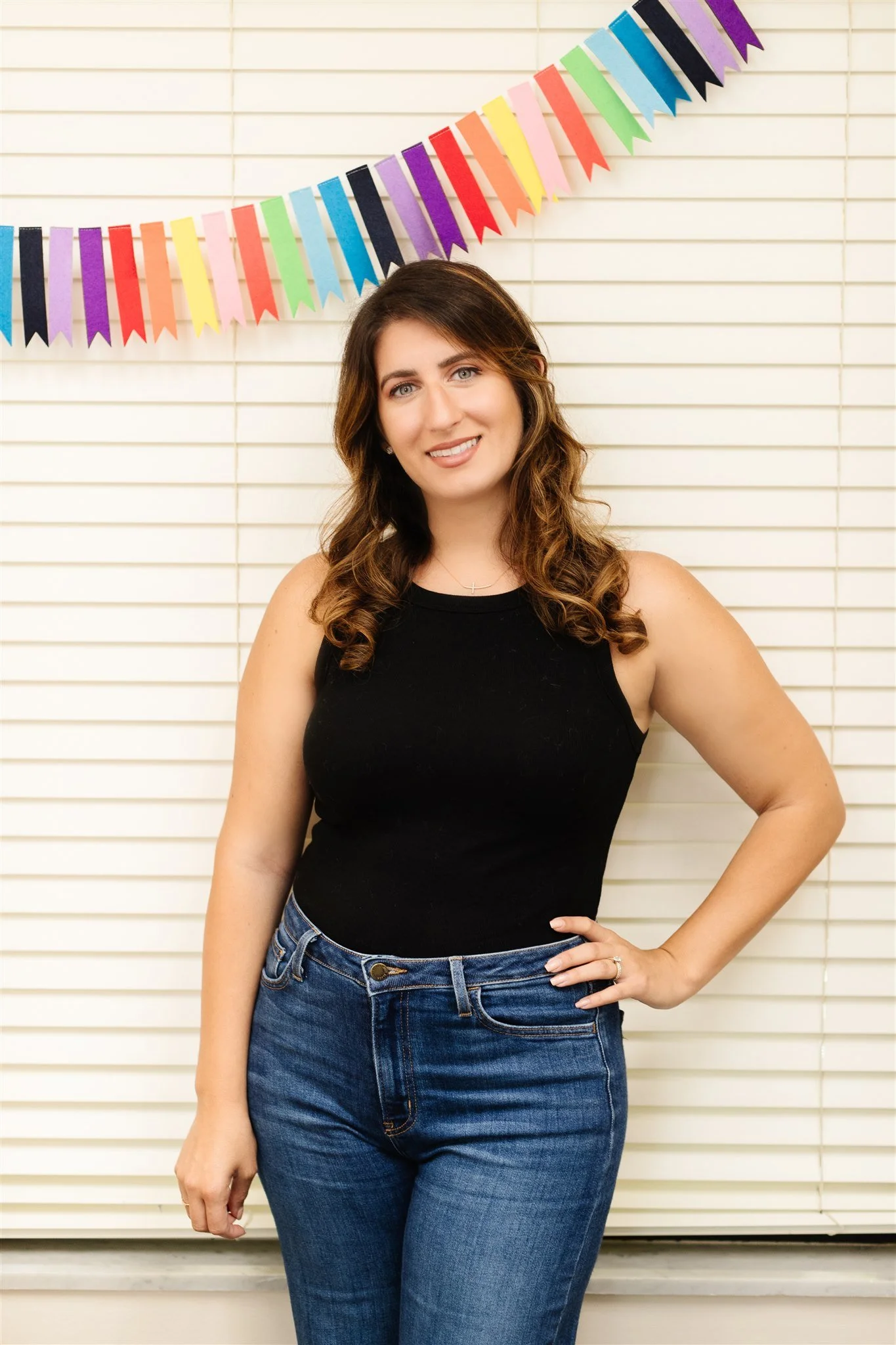 A young woman with long wavy brown hair, wearing a black sleeveless top and blue jeans, standing in front of white window blinds and colorful bunting.